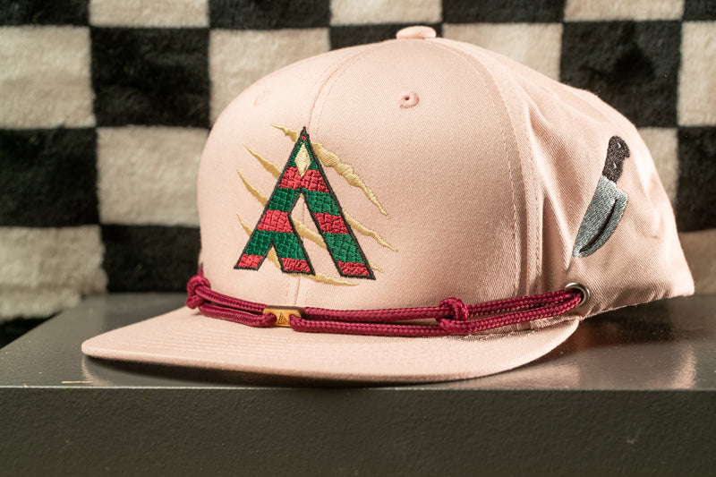 A pink baseball cap with a red and green embroidered logo on the front, sitting on a black surface against a checkered background.