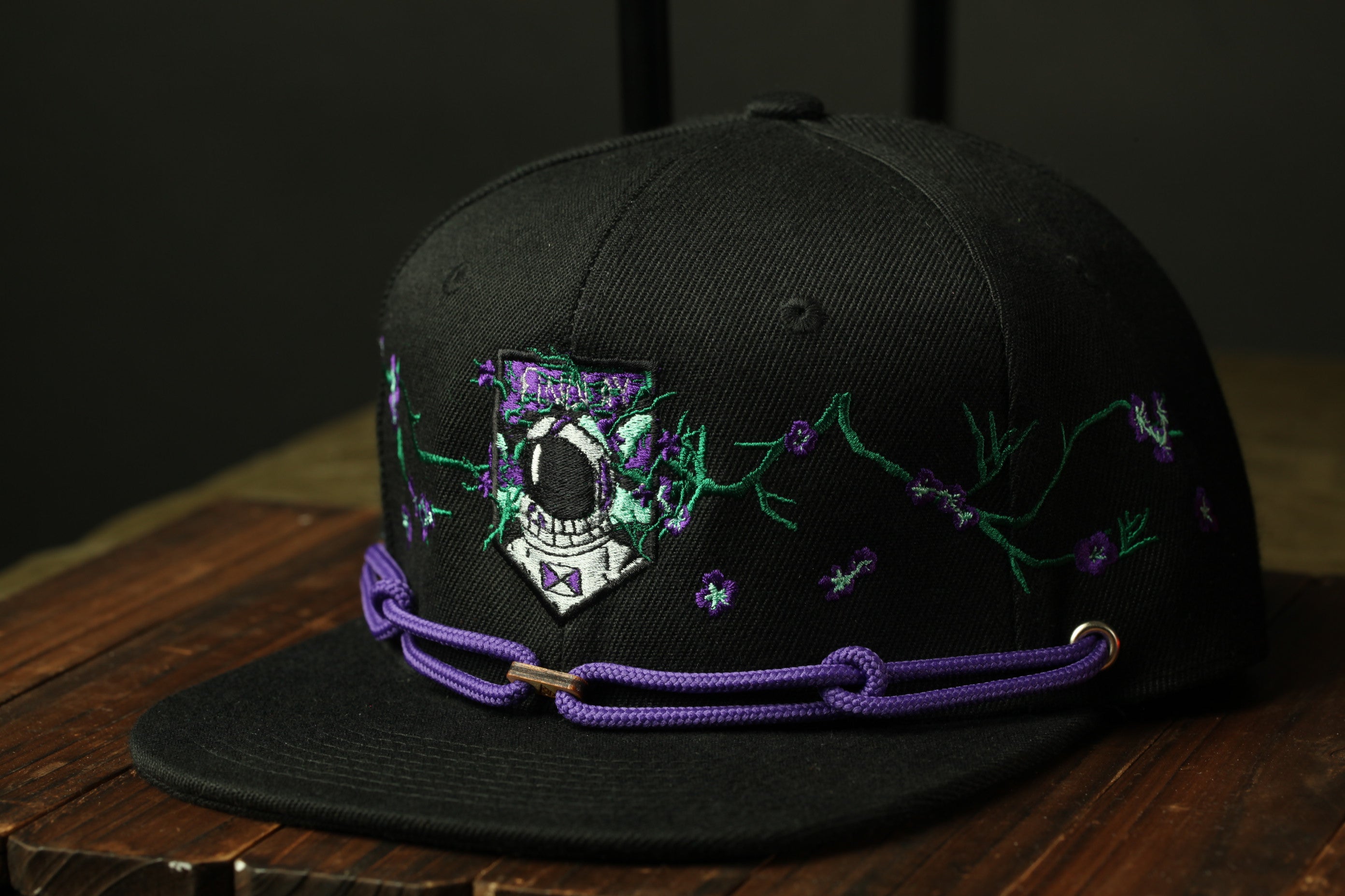 A black baseball cap with purple and green embroidered designs, including a shield-shaped emblem, is displayed on a wooden surface.