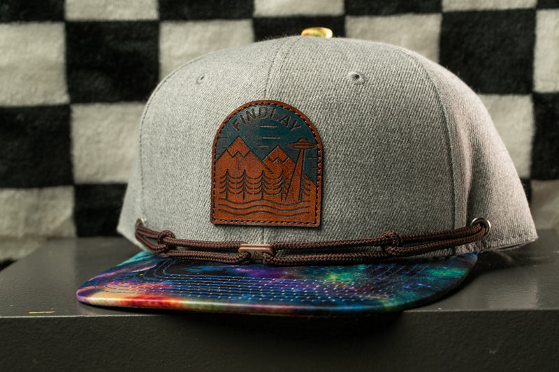 A gray baseball cap with a leather patch featuring a mountain landscape and a small alien figure, placed on a black surface against a checkered background.