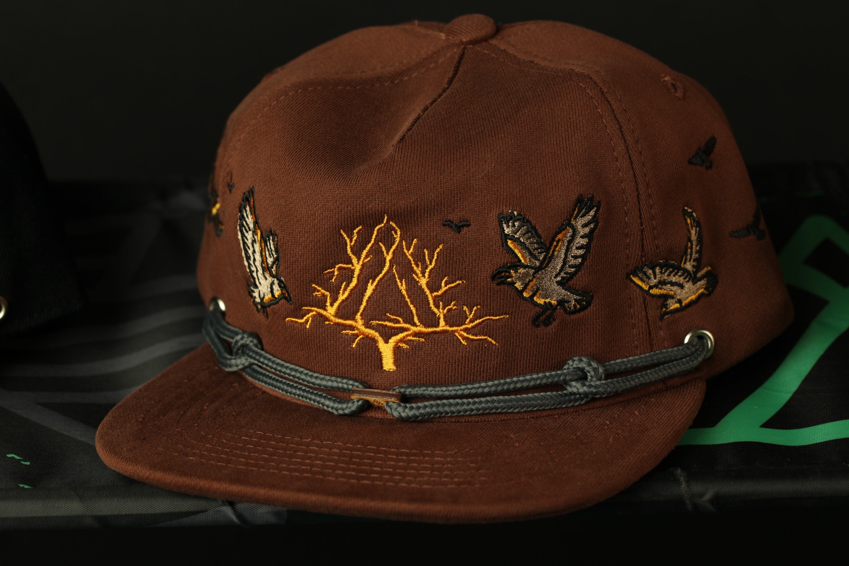 A brown baseball cap with a tree design and birds on it, resting on a black surface.