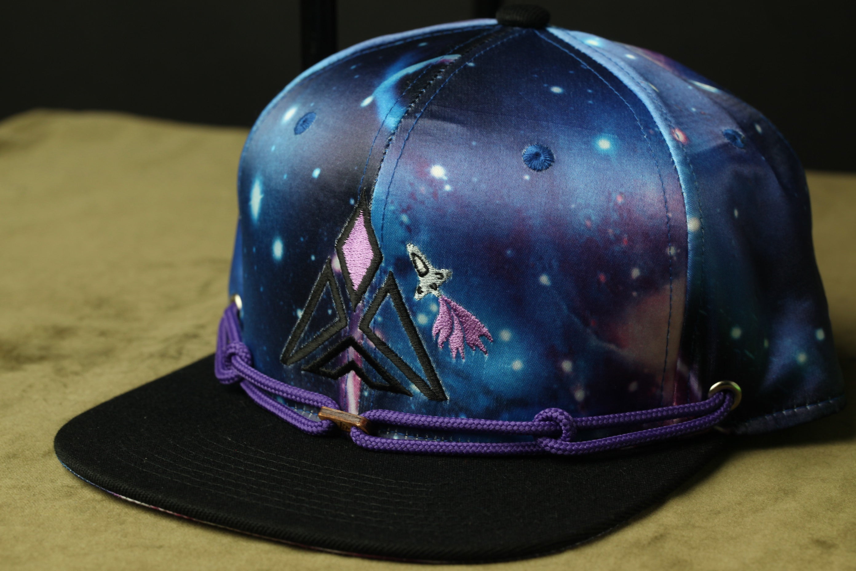 A colorful, patterned baseball cap with a space-themed design, featuring a purple and black geometric pattern and a small purple bird-like figure.