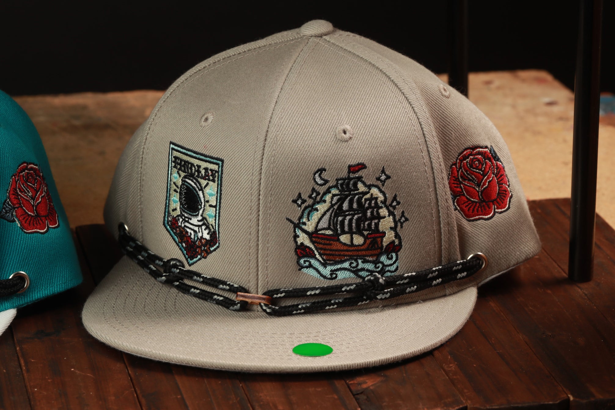 A gray baseball cap with various embroidered designs, including a ship, a rose, and a space-themed motif, is displayed on a wooden surface.
