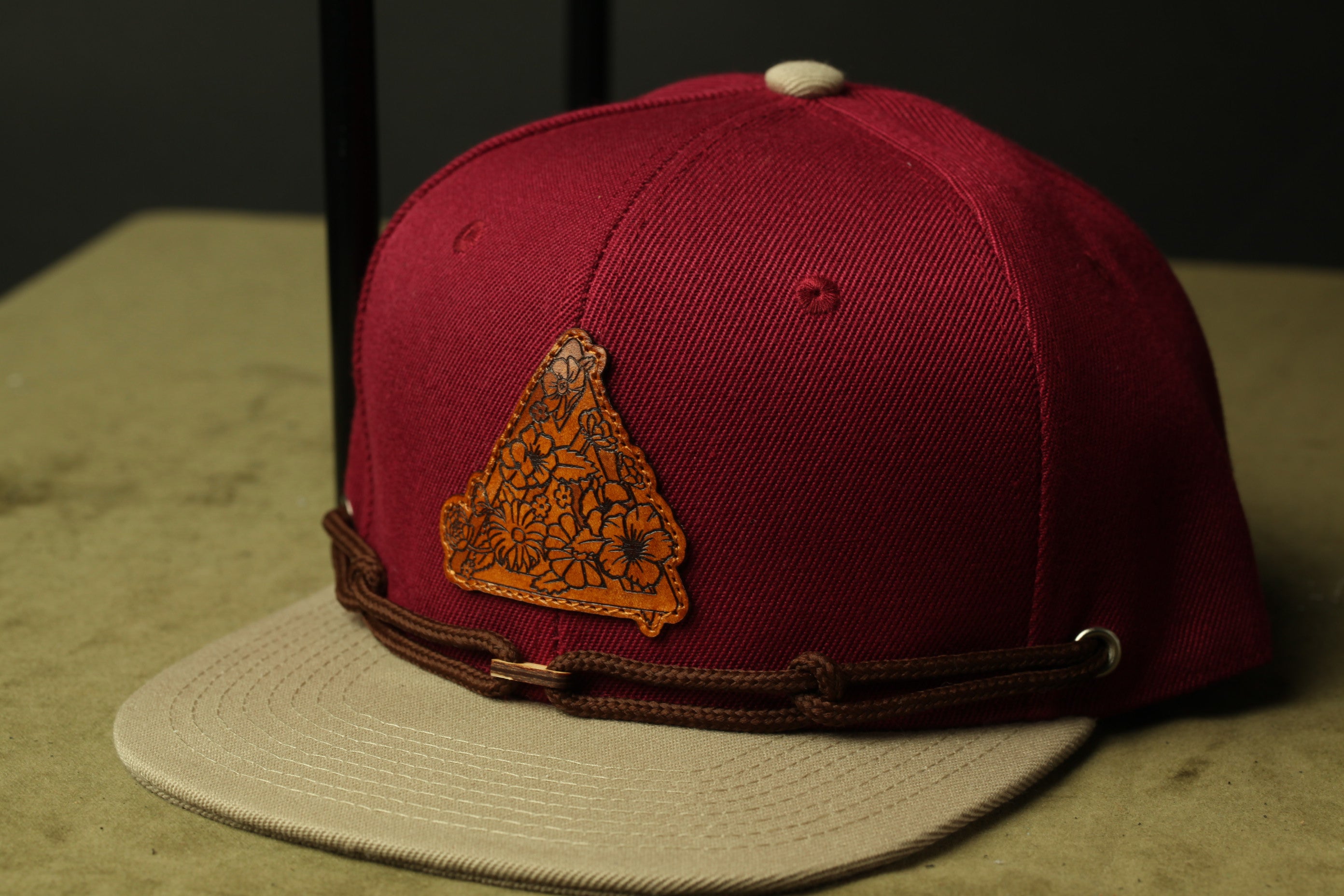 A maroon baseball cap with a brown leather patch featuring a floral design, placed on a beige surface.
