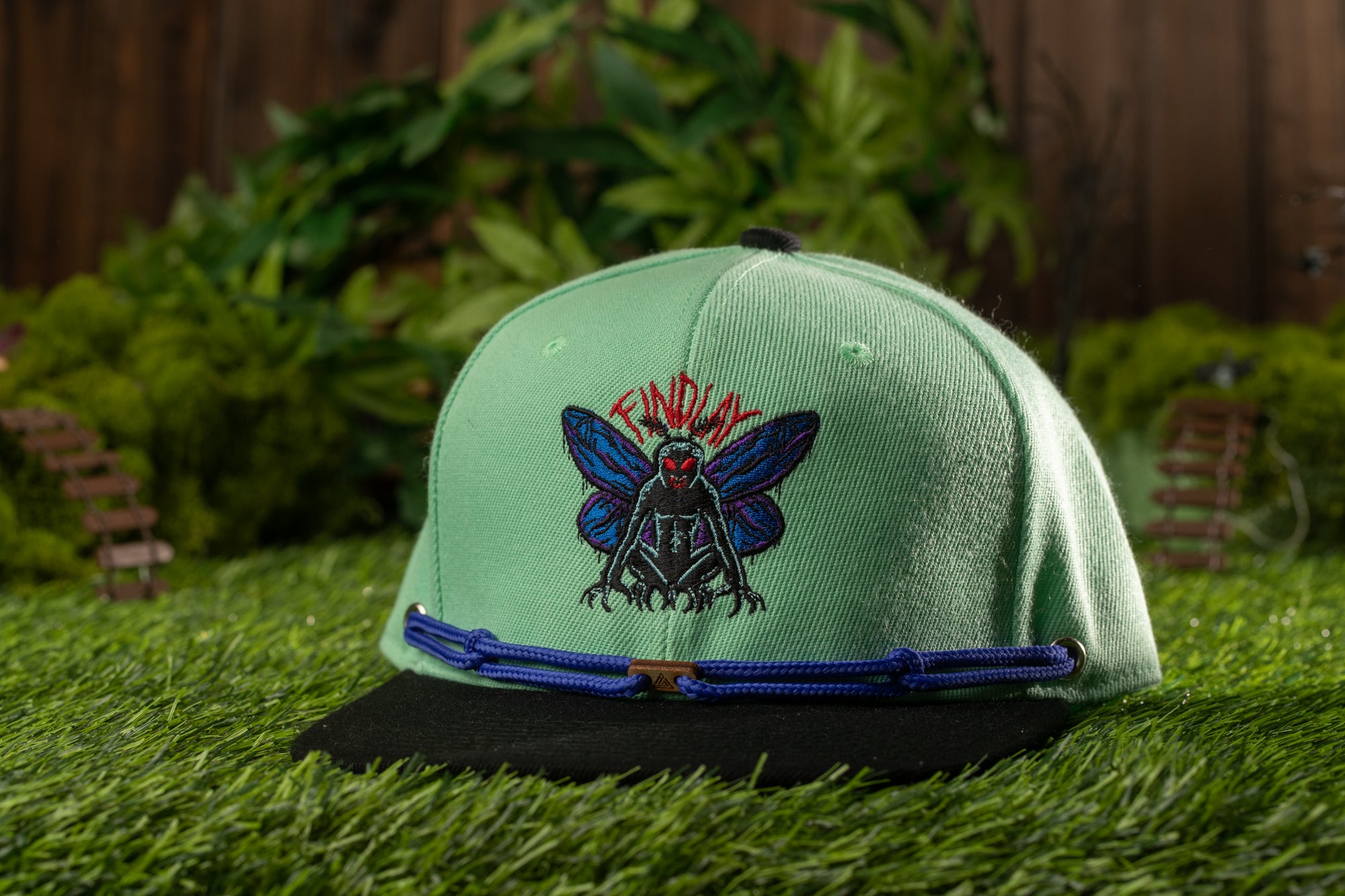 A green baseball cap with a blue and red design featuring a large insect-like creature, placed on a grassy surface with a wooden fence and greenery in the background.