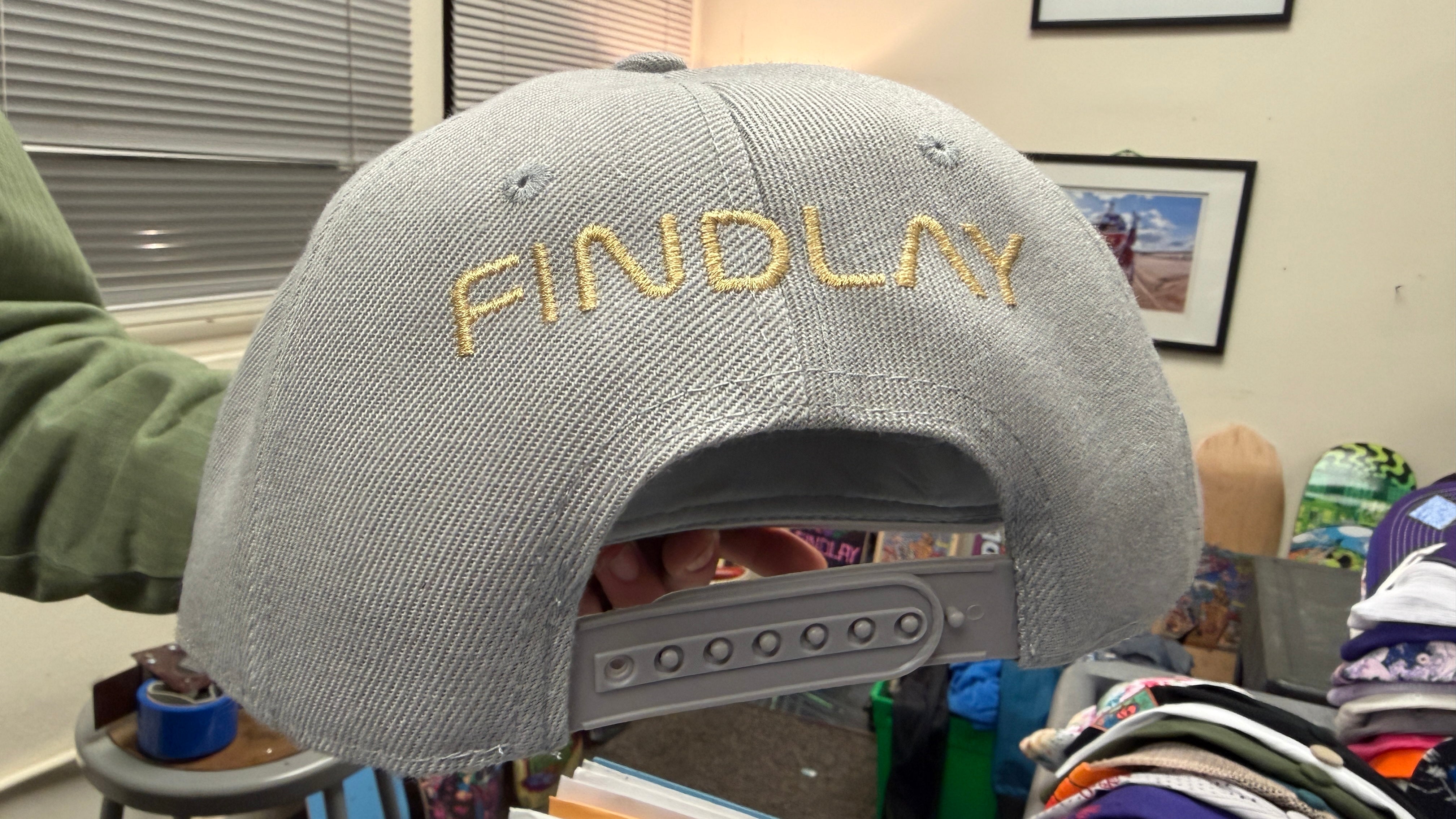 a gray baseball cap with the word "FINDlay" embroidered on it, displayed in a room with various other items visible in the background.