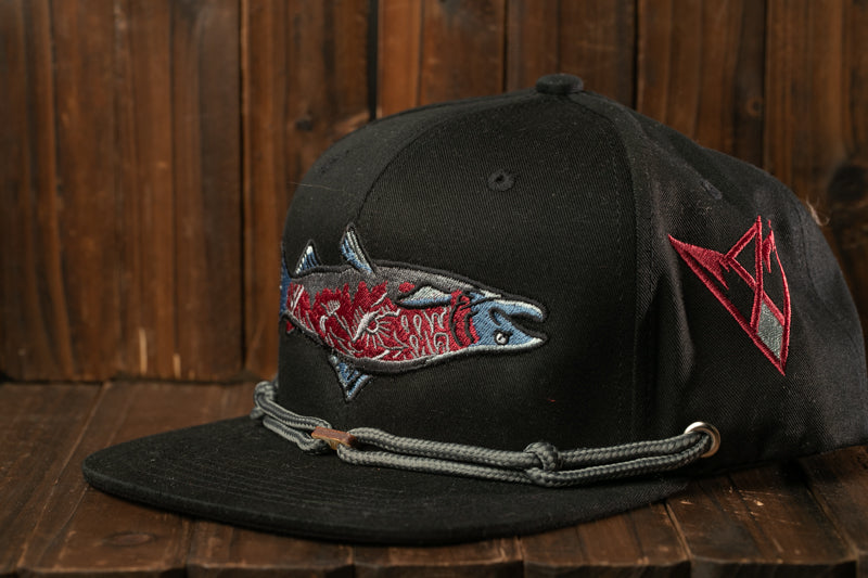 A black baseball cap with a fish embroidered on it, sitting on a wooden surface.