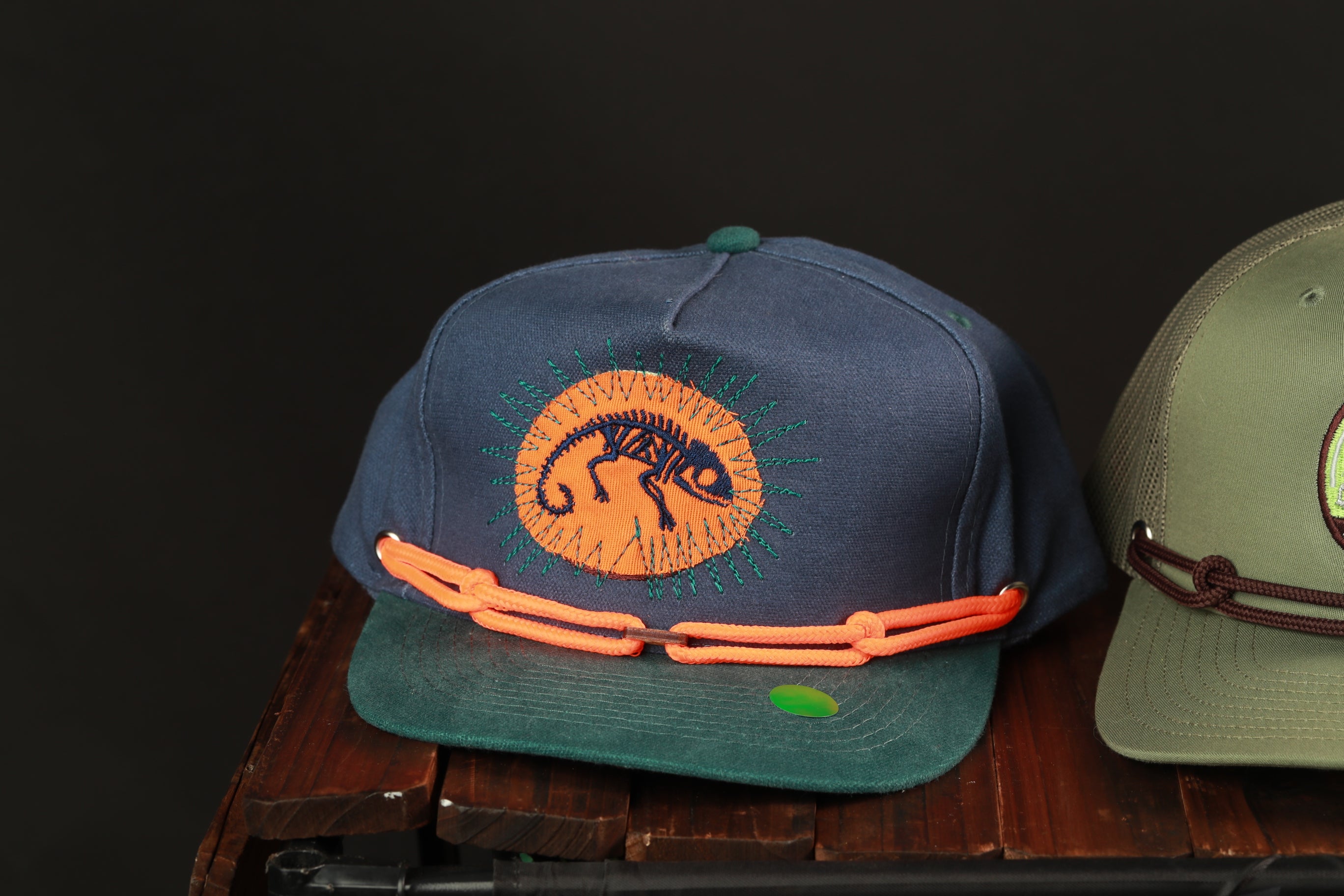 Two baseball caps are displayed on a wooden surface, one with a chameleon design and the other with a green and brown color scheme.