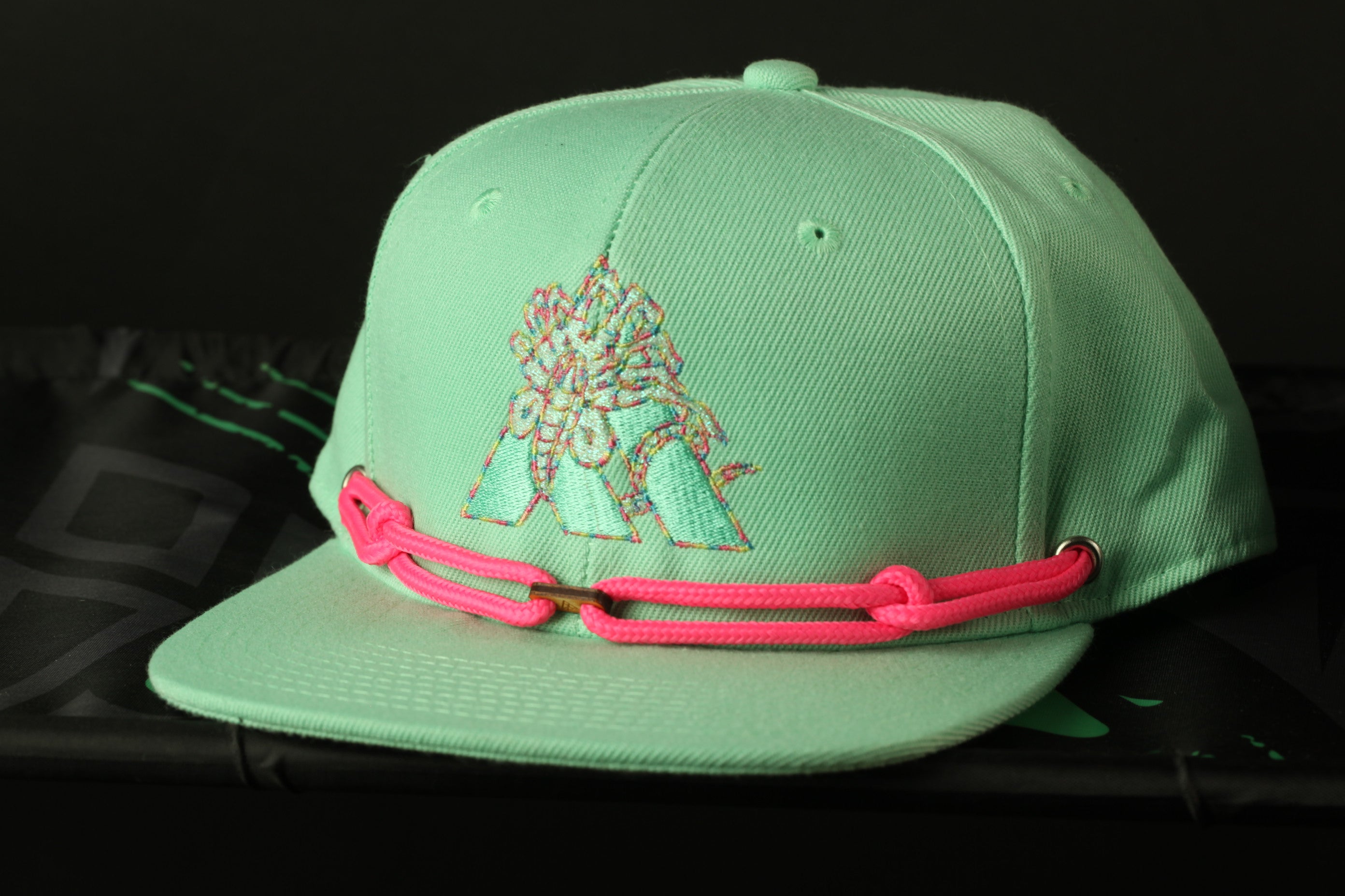 A mint green baseball cap with a pink chain strap is displayed against a black background.