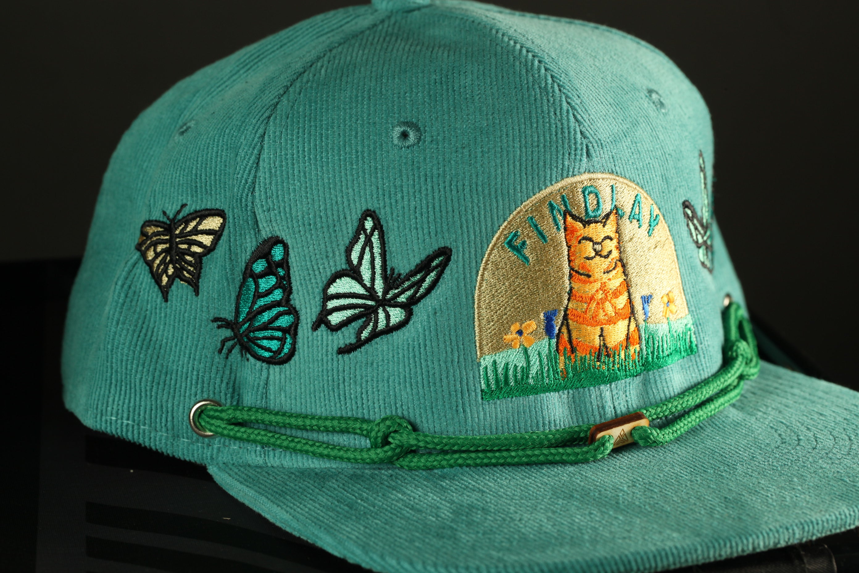 A turquoise baseball cap with various embroidered designs, including butterflies and a cat, and a small logo on the front.