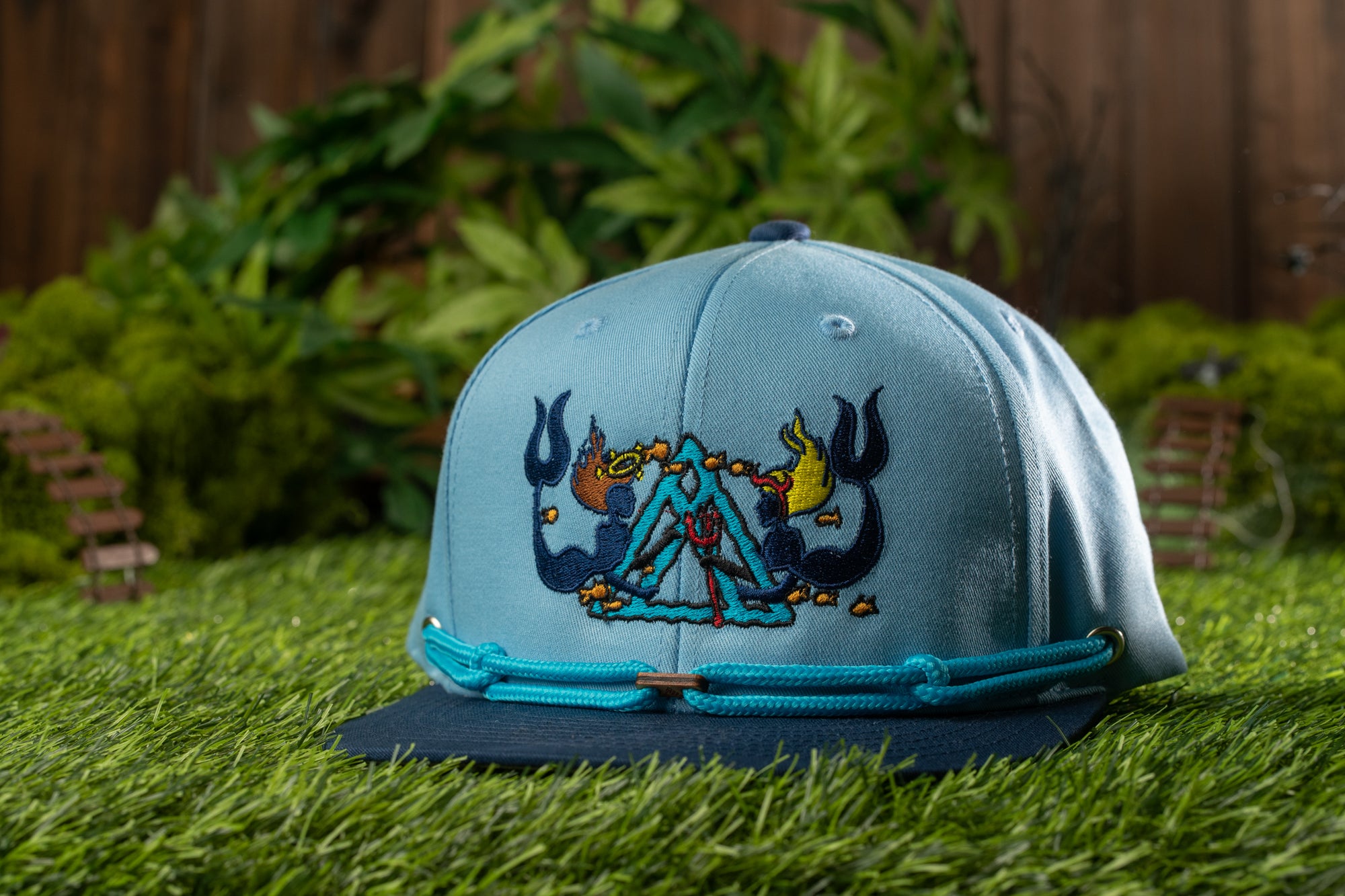 A blue baseball cap with a colorful, embroidered design sits on a grassy surface, with a wooden fence and greenery in the background.