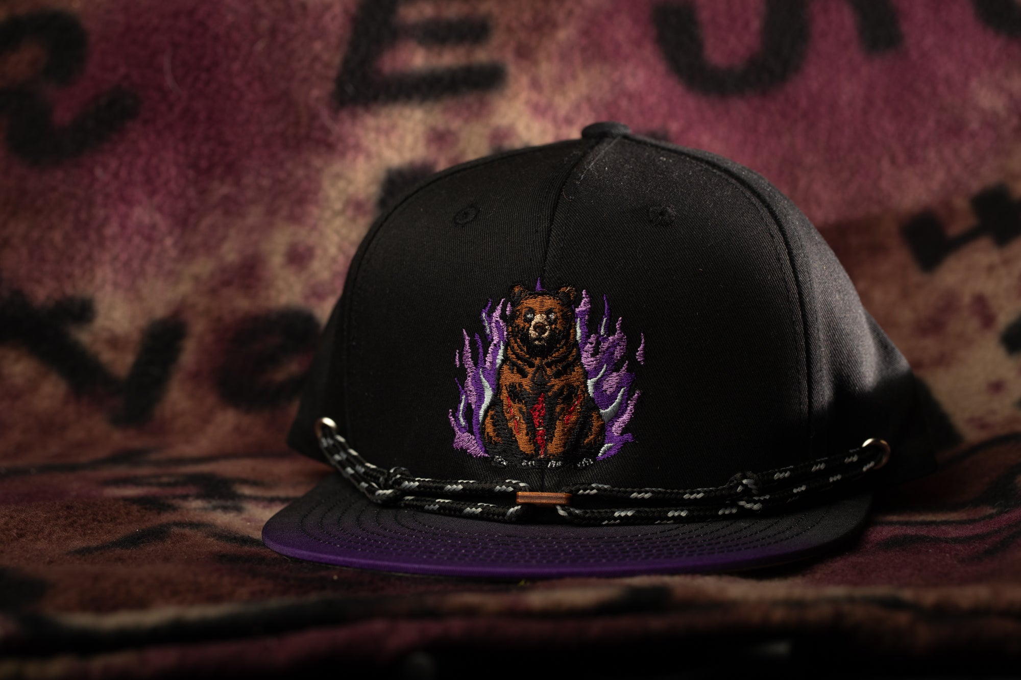 A black baseball cap with a purple brim and a bear embroidered on it, sitting on a patterned fabric.