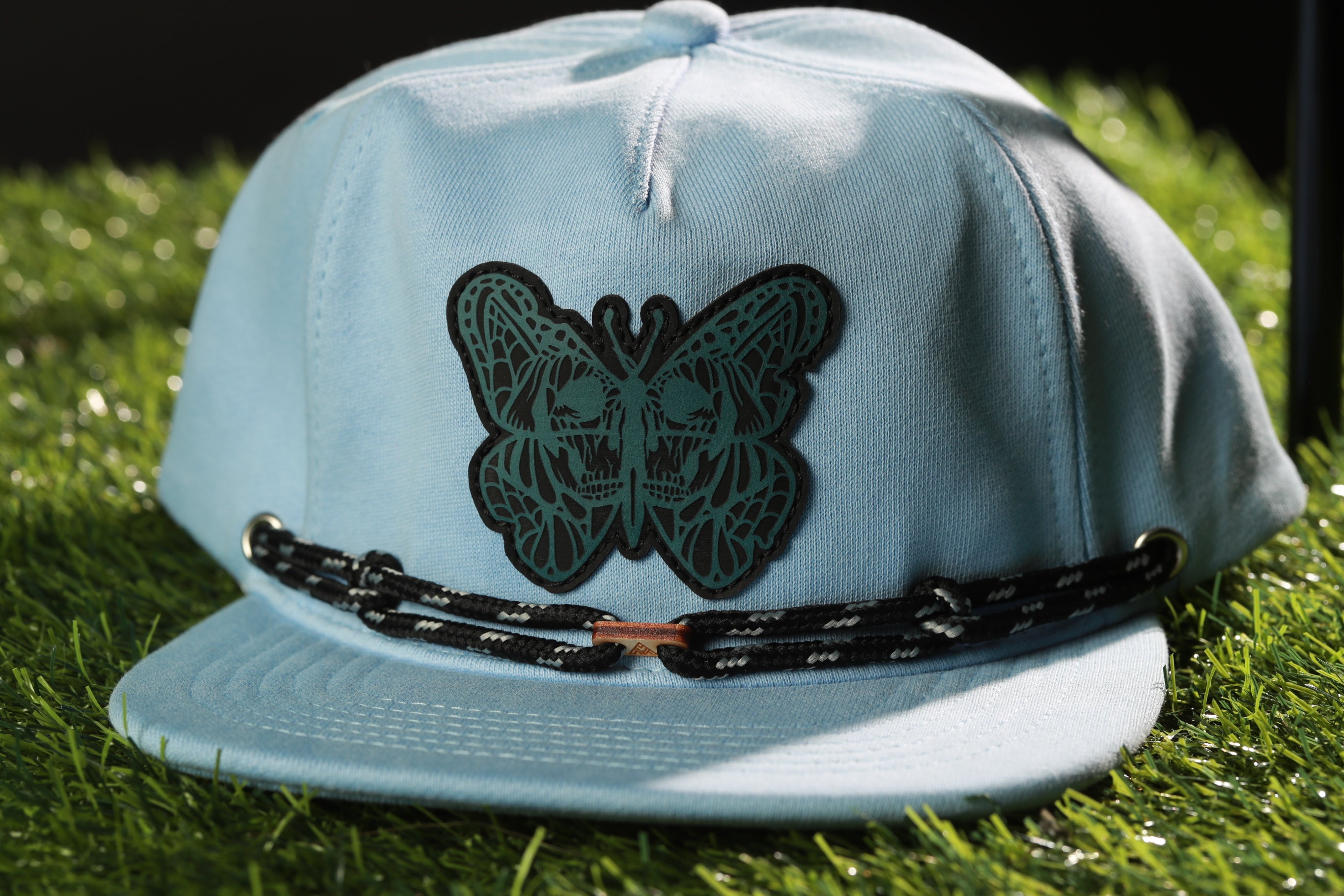 A light blue baseball cap with a black butterfly design on the front, resting on a grassy surface.