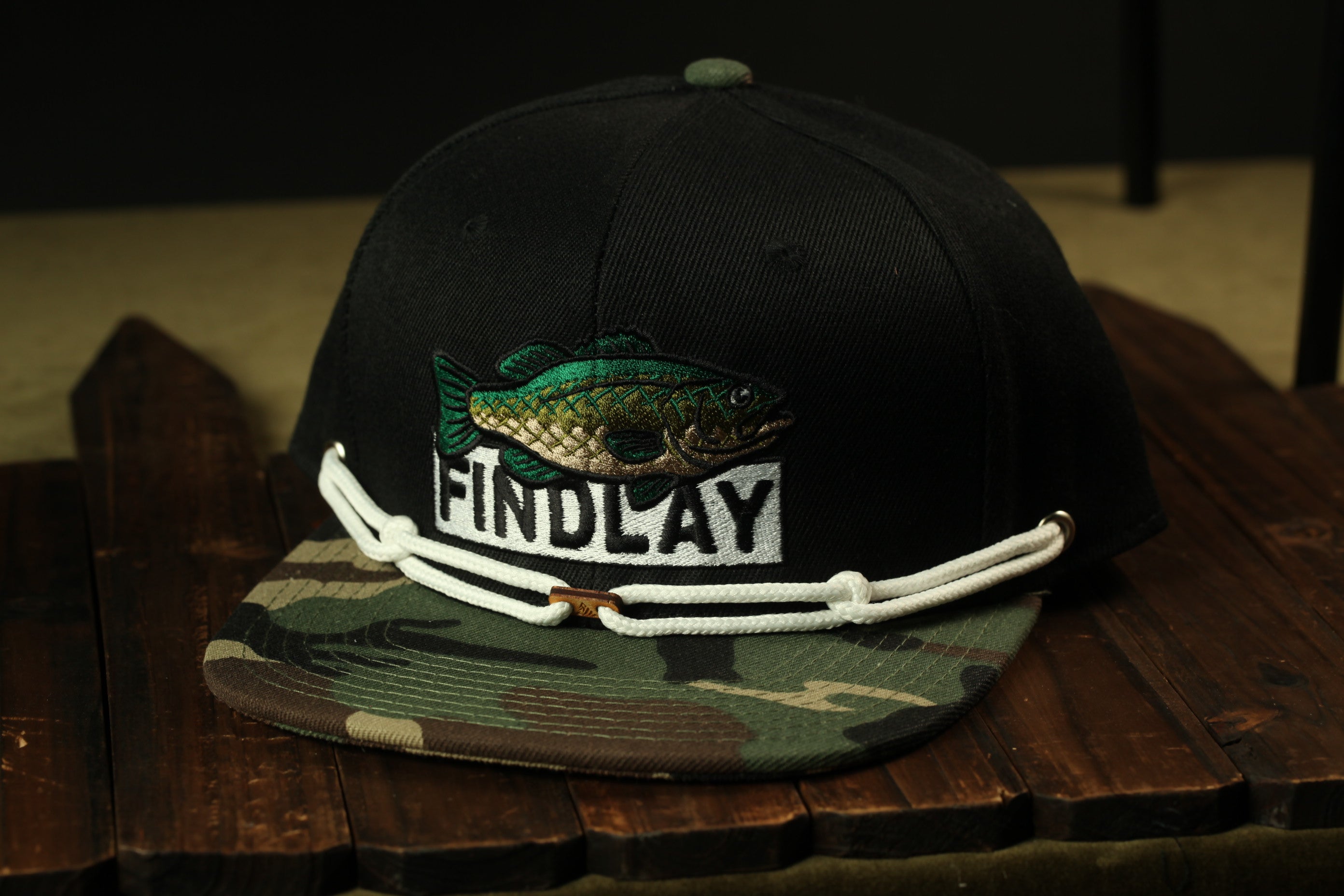 A black baseball cap with a camouflage pattern and a fish embroidered on it, sitting on a wooden surface.