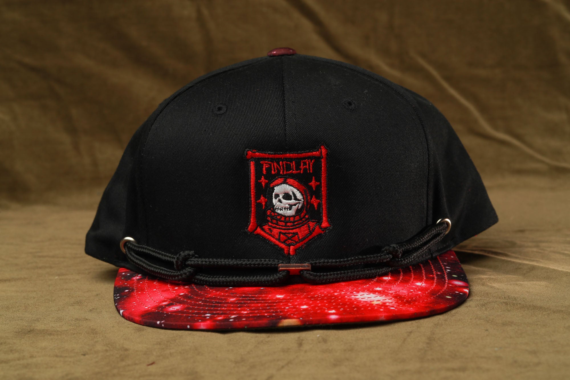 A black baseball cap with a red and black skull and crossbones design on the front, placed on a brown surface.