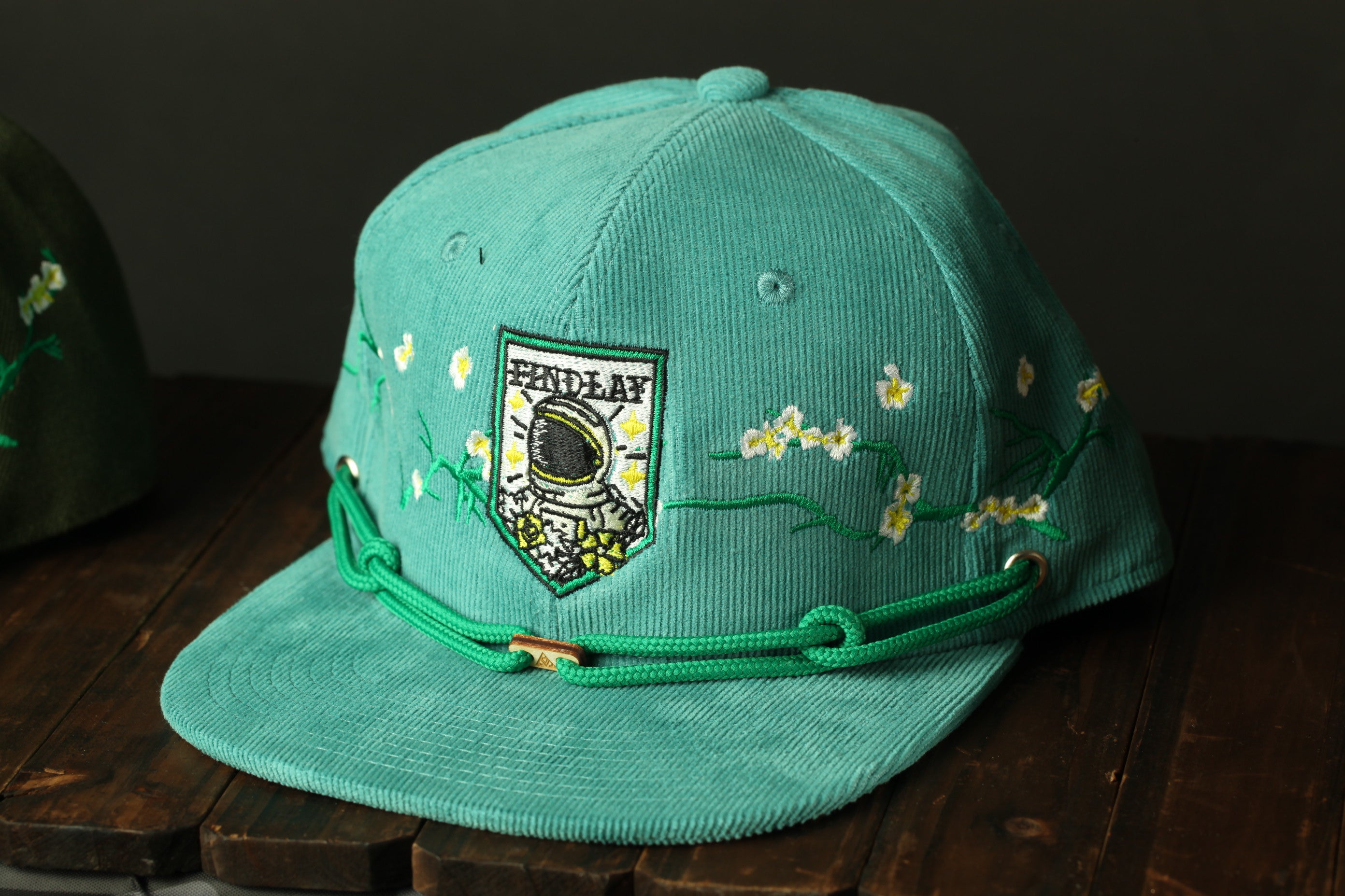 A turquoise baseball cap with a floral embroidered design and a small patch on the front, sitting on a wooden surface.