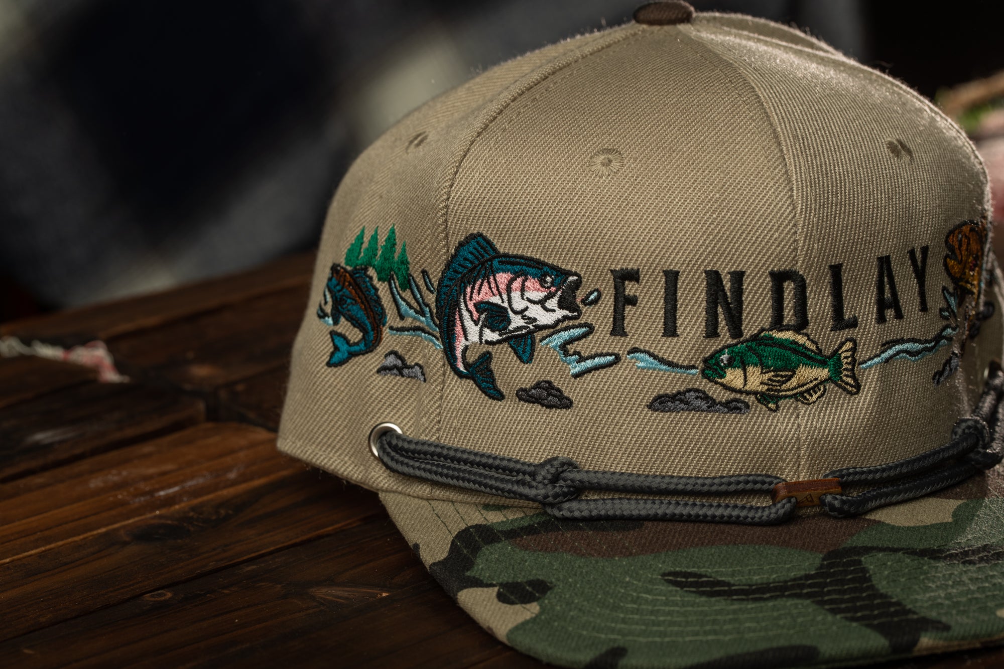 A beige baseball cap with a camouflage pattern and embroidered fish designs on the front.