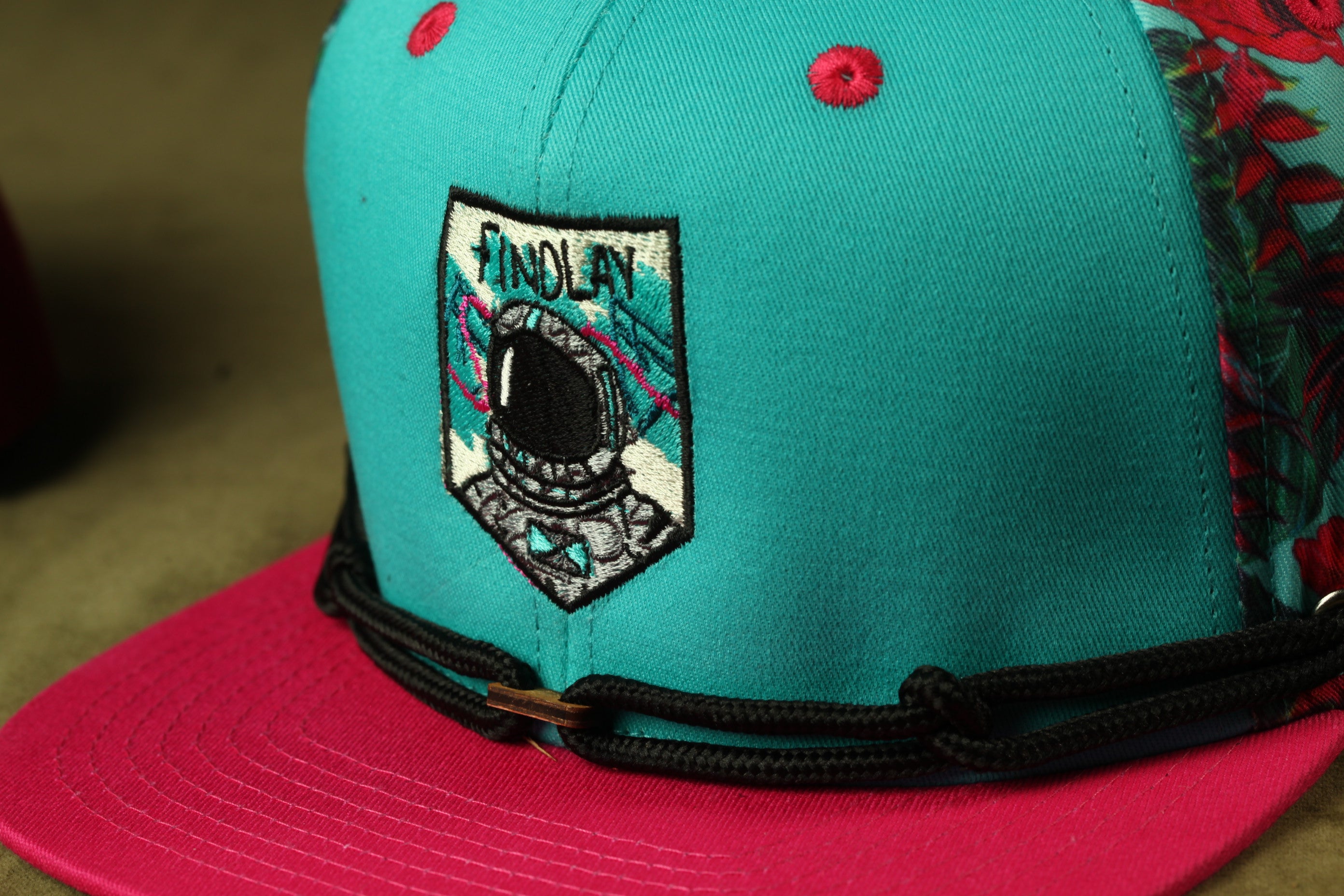 A close-up of a turquoise baseball cap with a black strap and a small embroidered patch on the front.