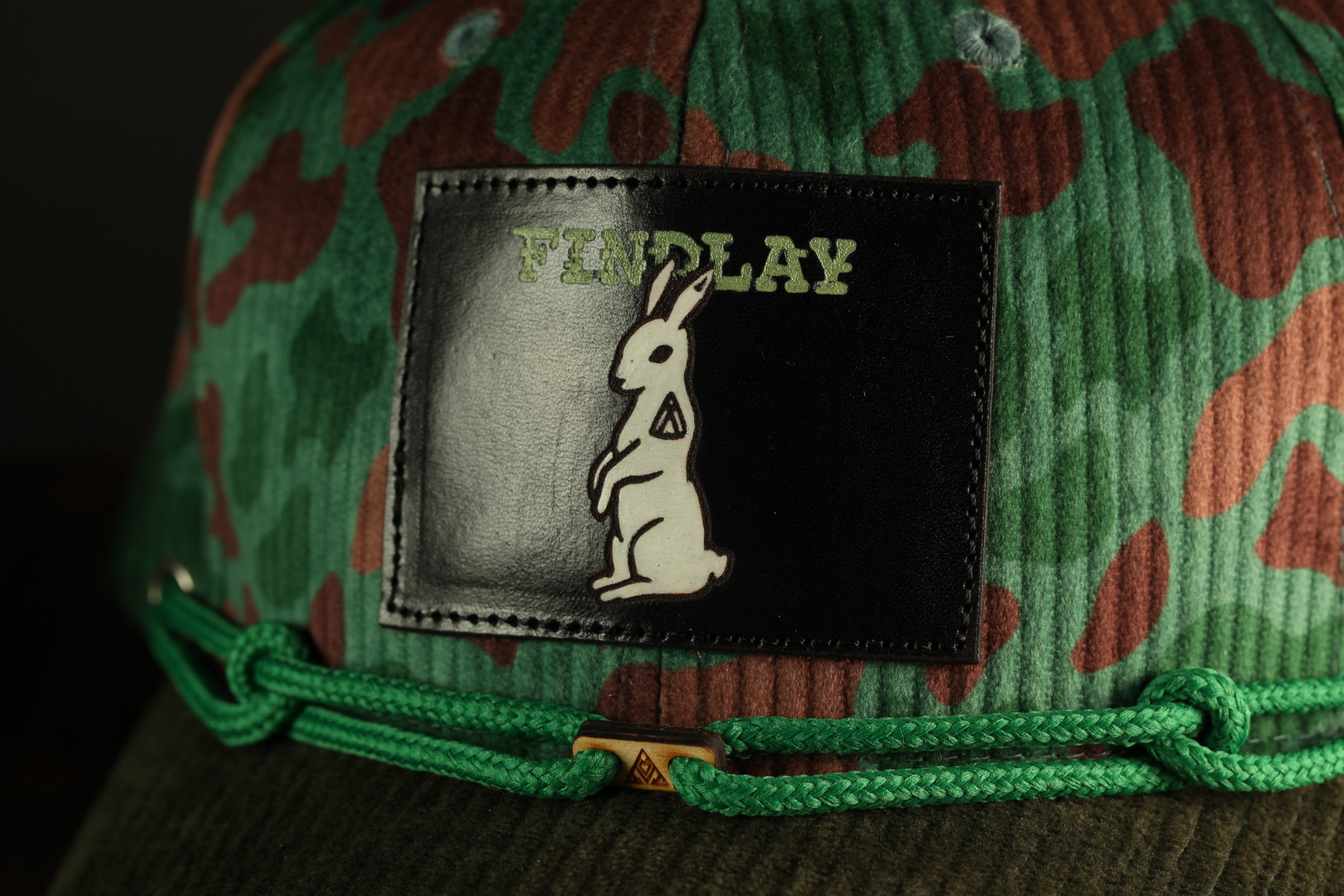 A close-up of a camouflage-patterned baseball cap with a patch featuring a cartoon rabbit and the word "FINDA" printed on it.
