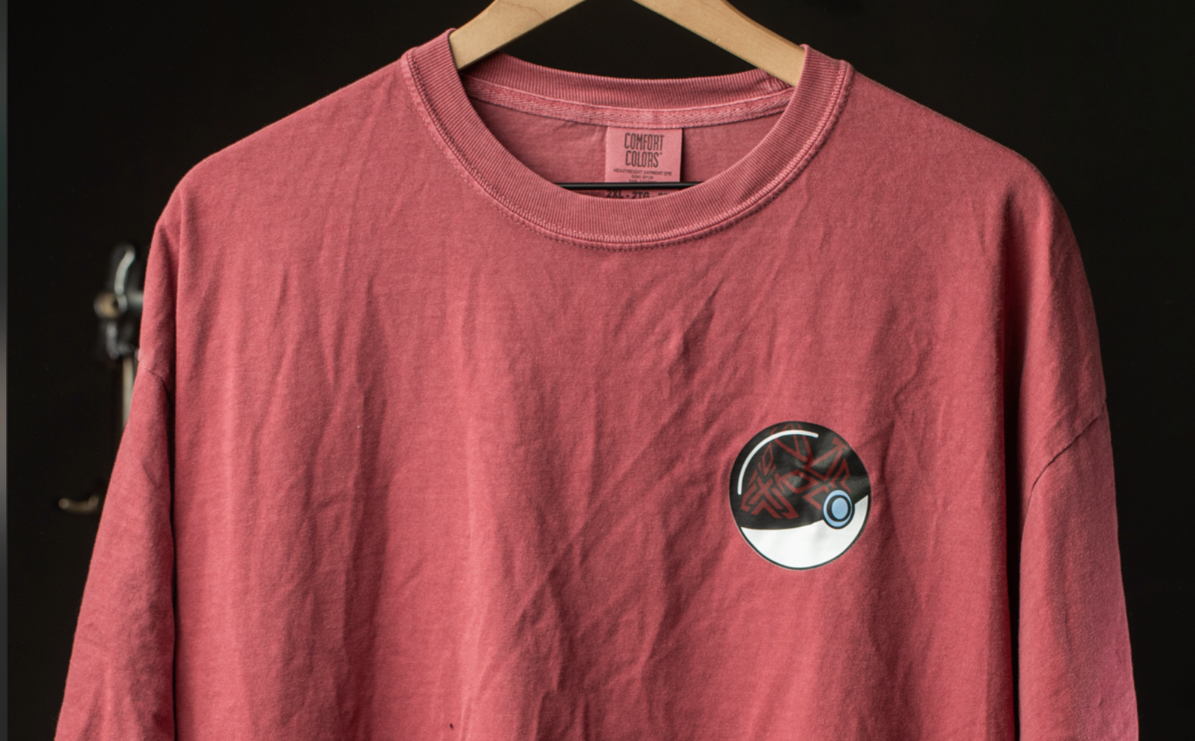 A red t-shirt with a graphic design on the front, hanging on a wooden hanger against a dark background.