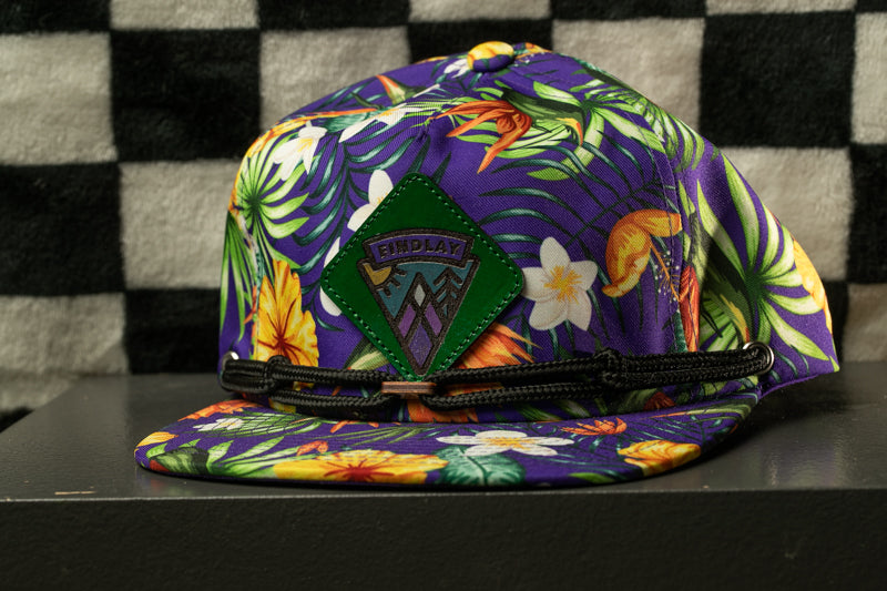 A colorful, patterned baseball cap with a tropical print design, featuring a green patch with a mountain and palm leaves.