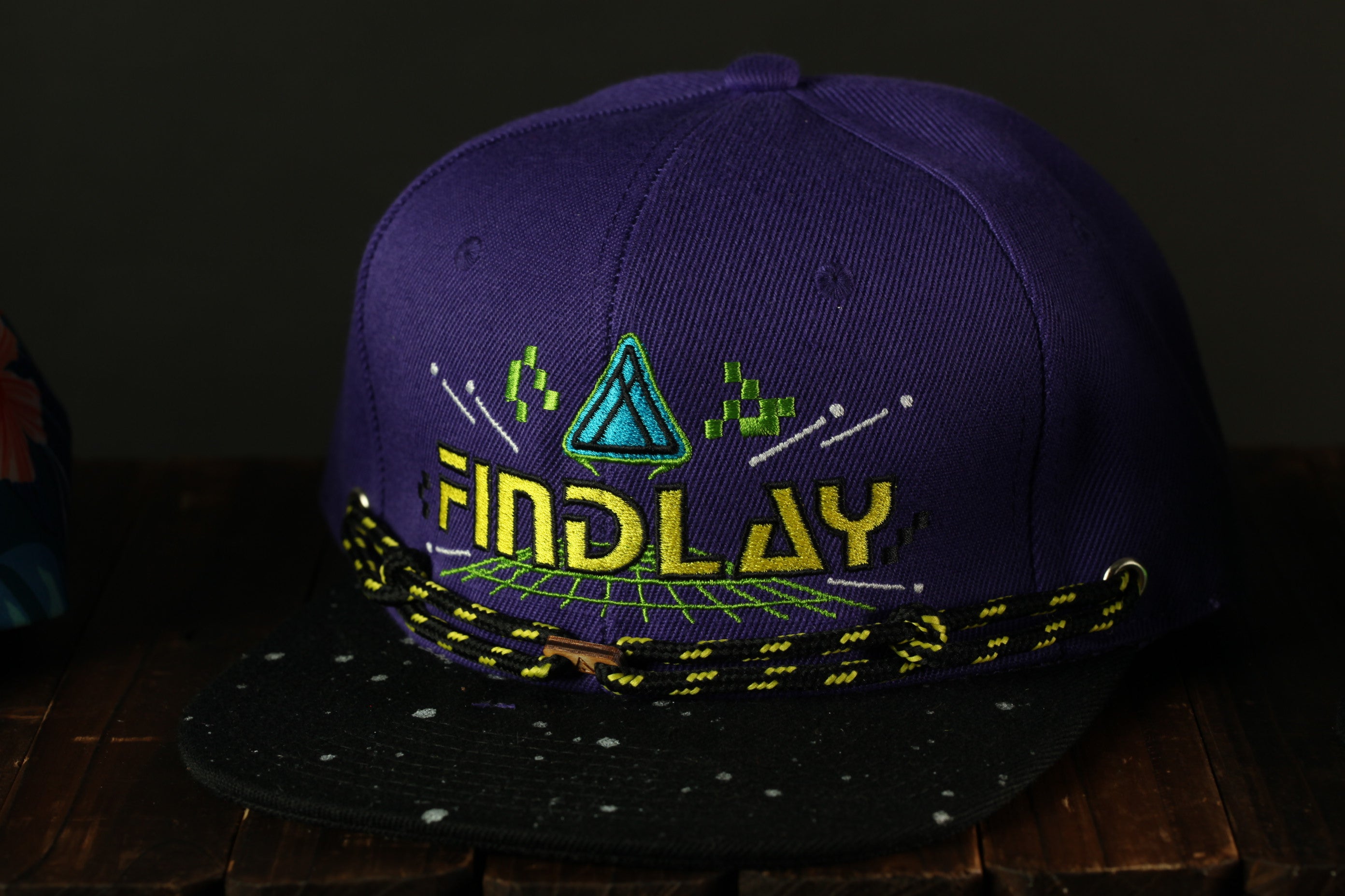 A purple baseball cap with a black brim and yellow and green embroidered designs, including a triangle and stars, is displayed on a wooden surface.