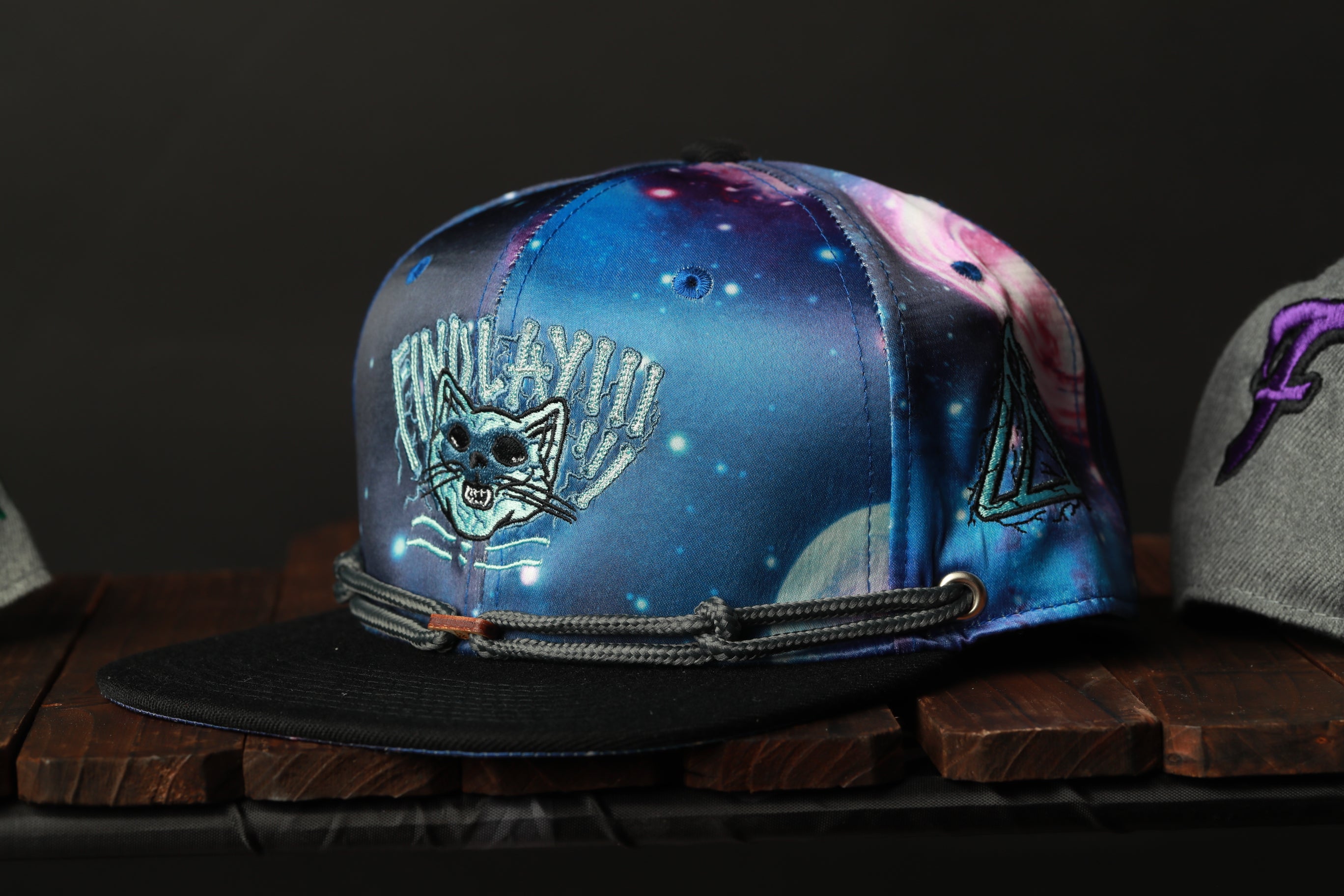 A colorful baseball cap with a cat design and a space-themed pattern, displayed on a wooden surface.