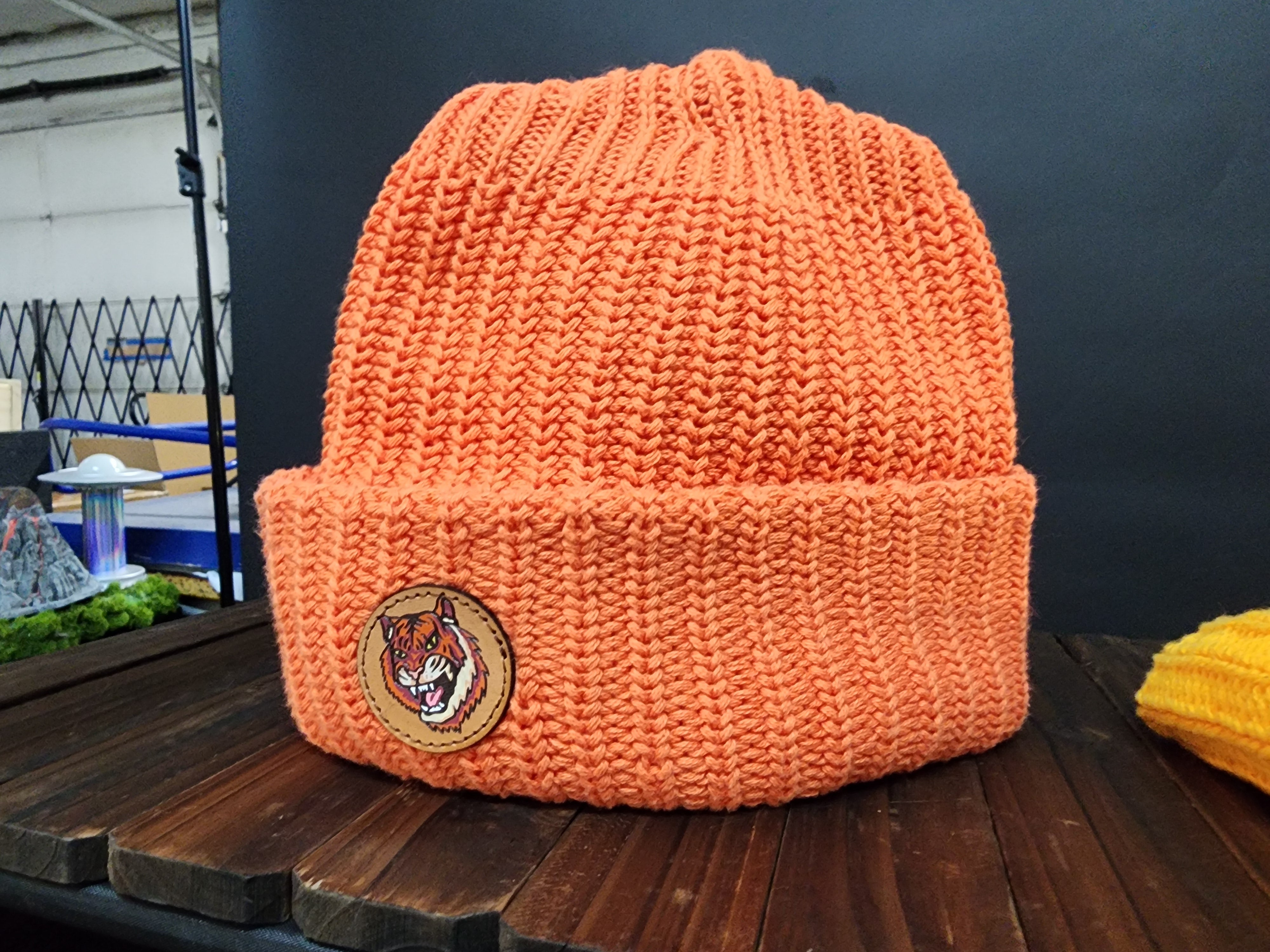An orange knitted beanie with a cat patch is displayed on a wooden surface.