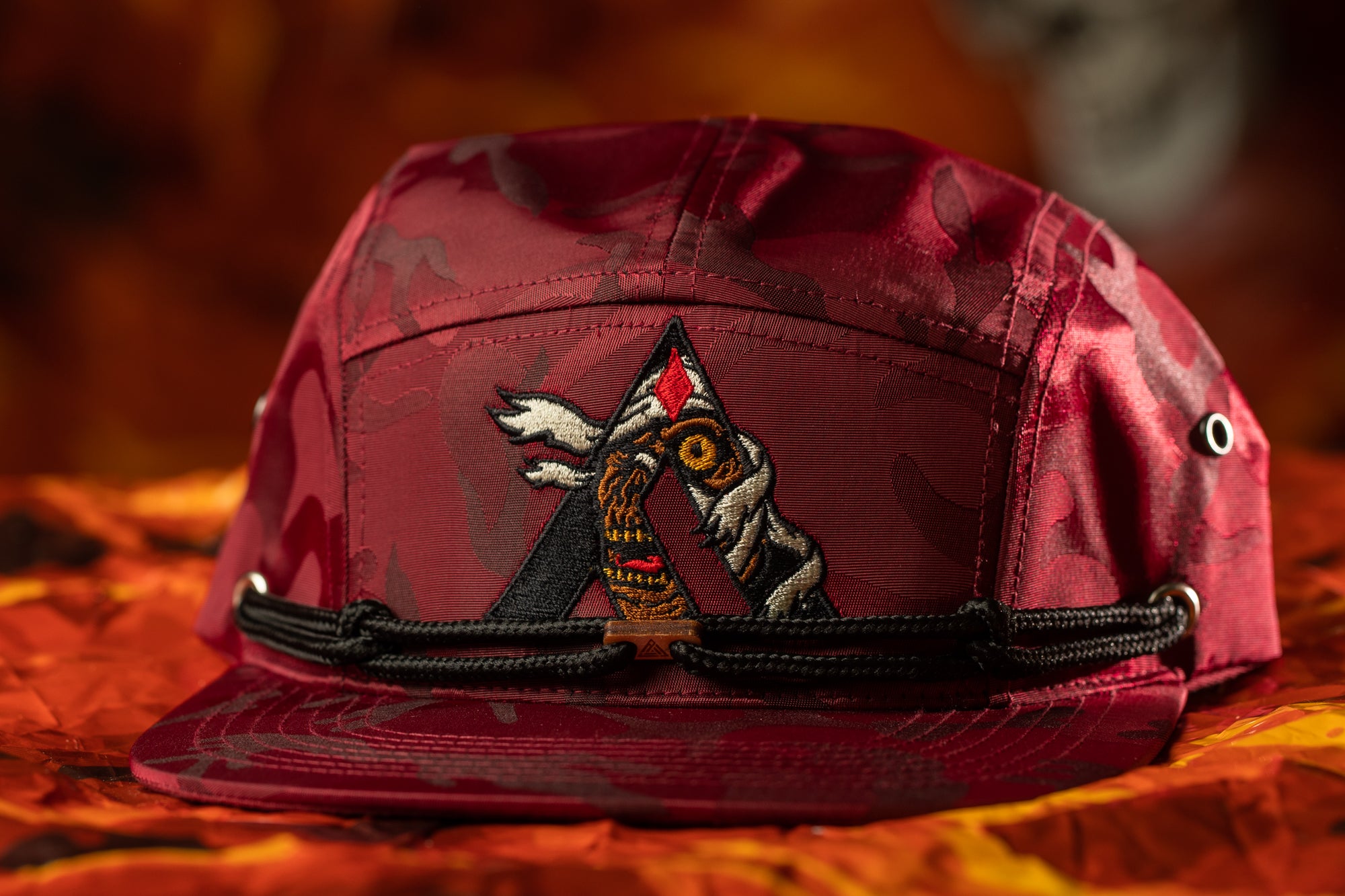 A red camouflage baseball cap with a black band and a decorative embroidered logo on the front.