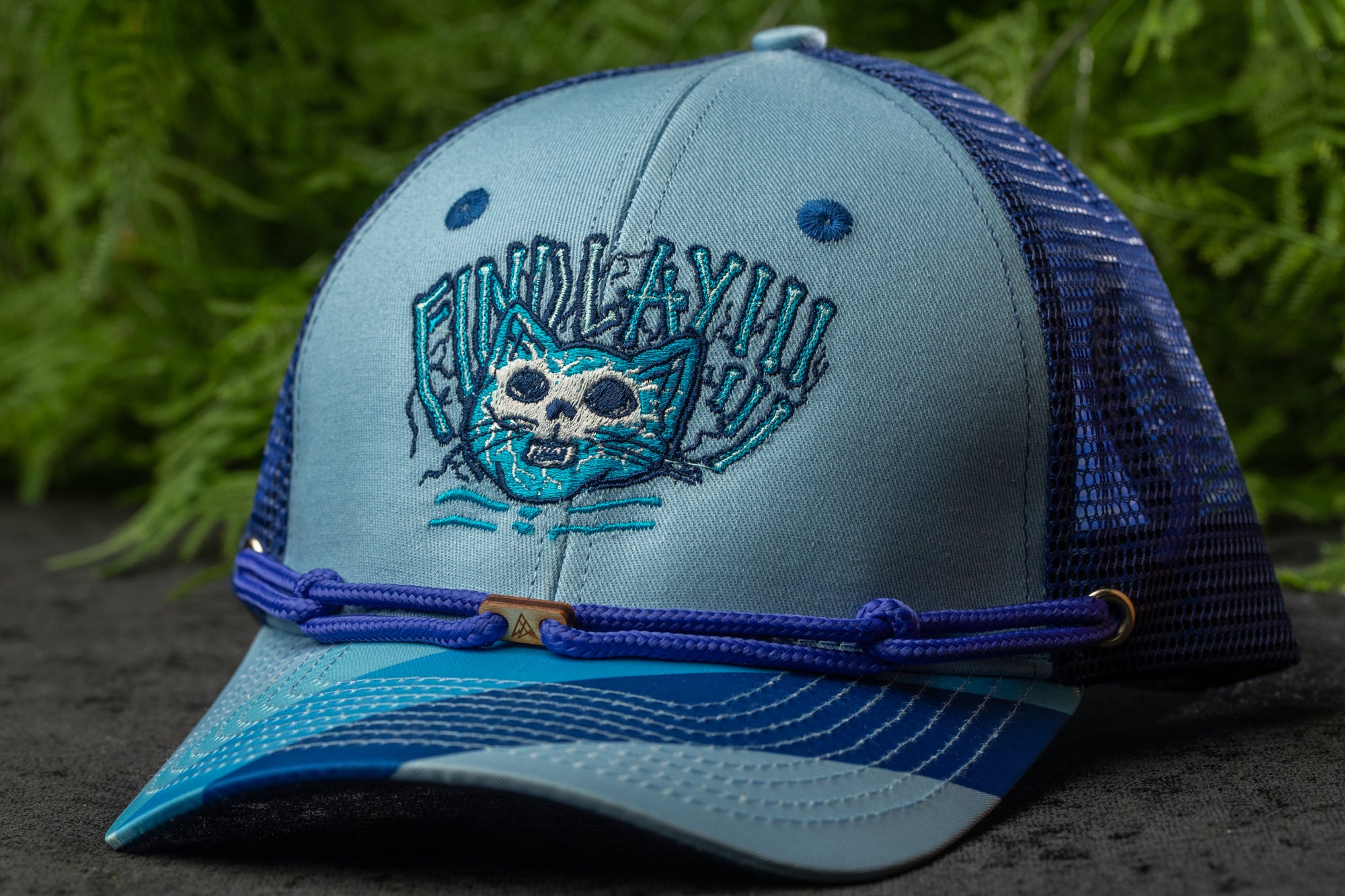 A blue and gray baseball cap with a cat embroidered on the front, sitting on a surface with green foliage in the background.