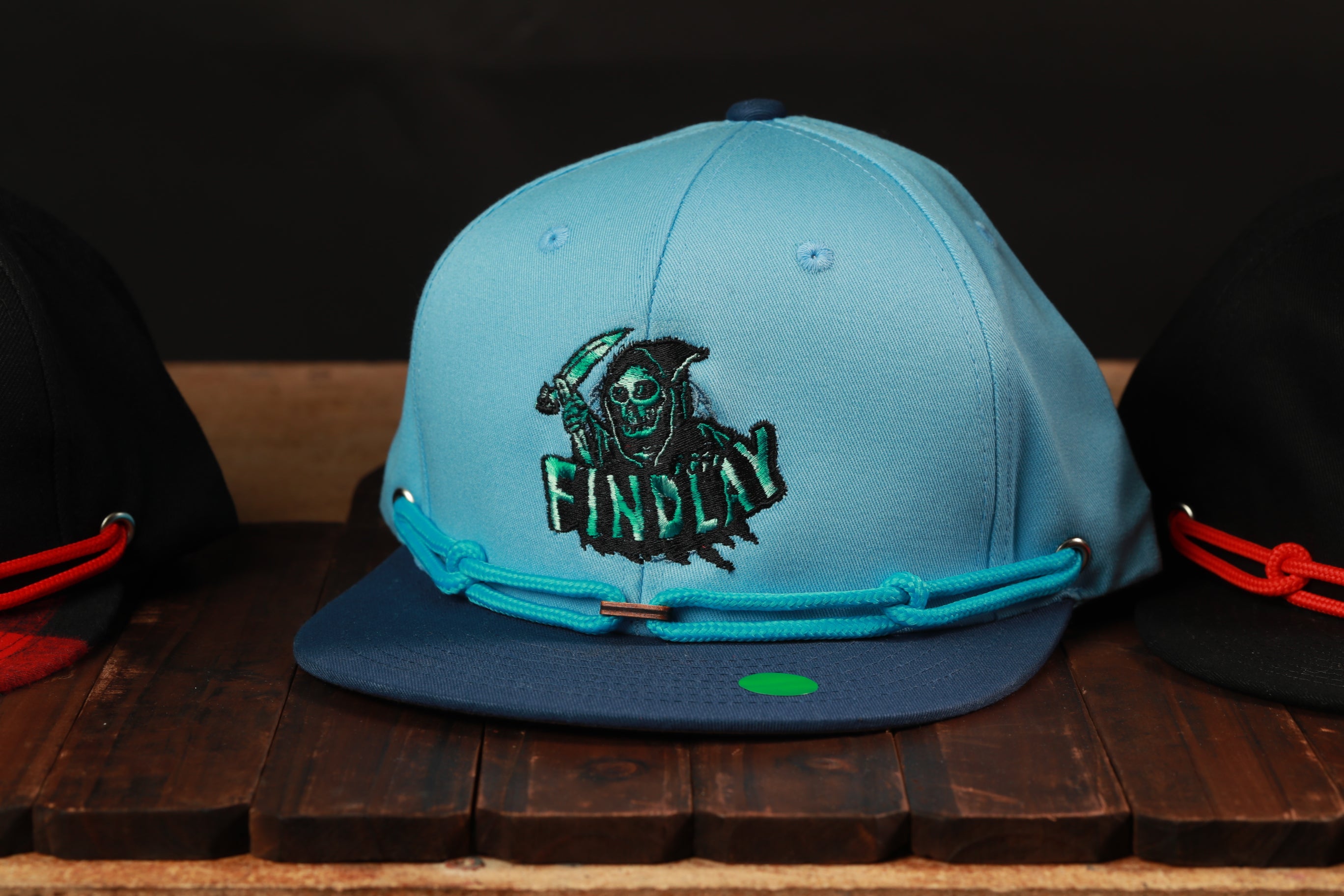 A blue baseball cap with a skull and crossbones design, featuring the word "FINDlay" on the front, is displayed on a wooden surface.