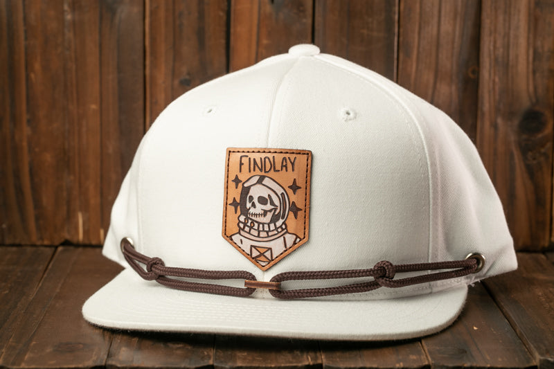A white baseball cap with a brown leather patch featuring a skull and the word "FINDLAY" on it, placed on a wooden surface.