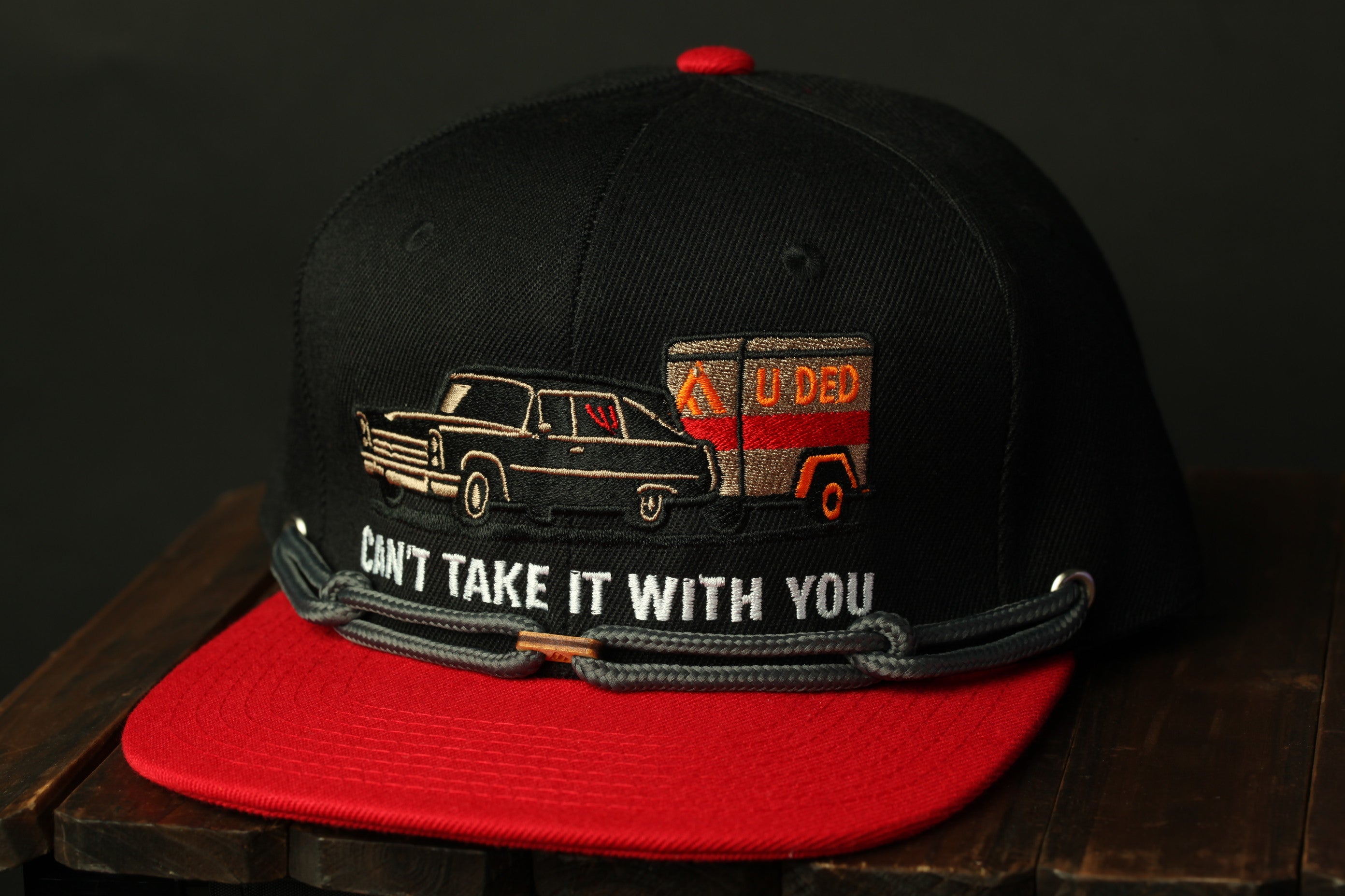 A black baseball cap with a red brim, featuring an embroidered design of a car and a trailer with the text "CANT TAKE IT WITH YOU" on it.