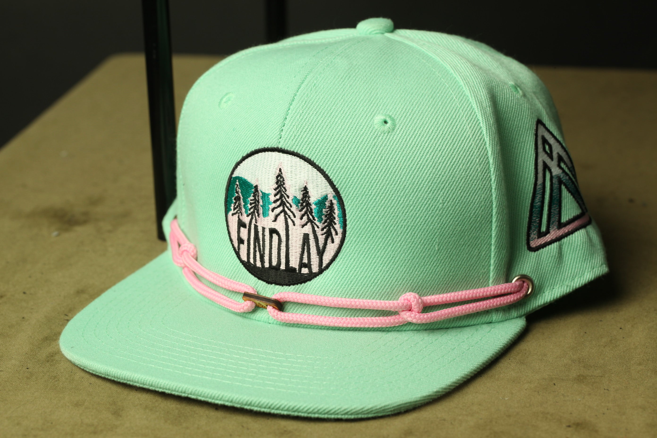 A mint green baseball cap with a pink rope and a circular logo on the front.