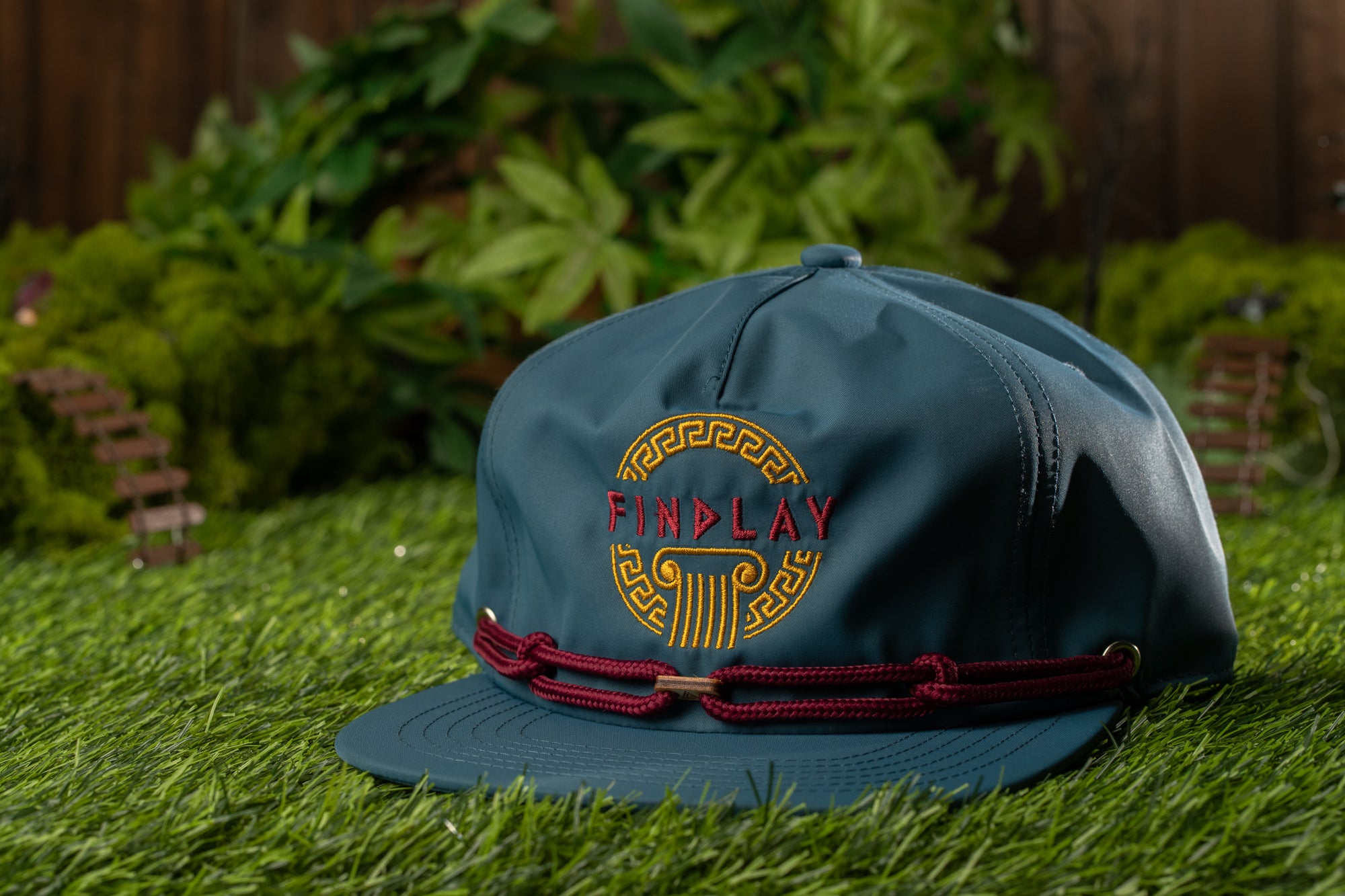 A blue baseball cap with a red band and the word "FINPLAY" printed on it, placed on a grassy surface with a blurred background of greenery.