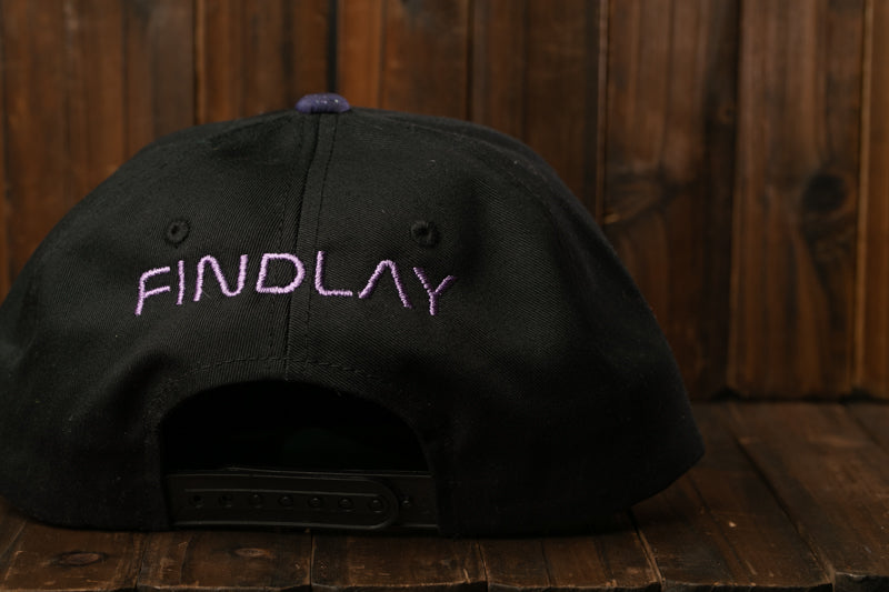 A black baseball cap with purple text on the front, sitting on a wooden surface.