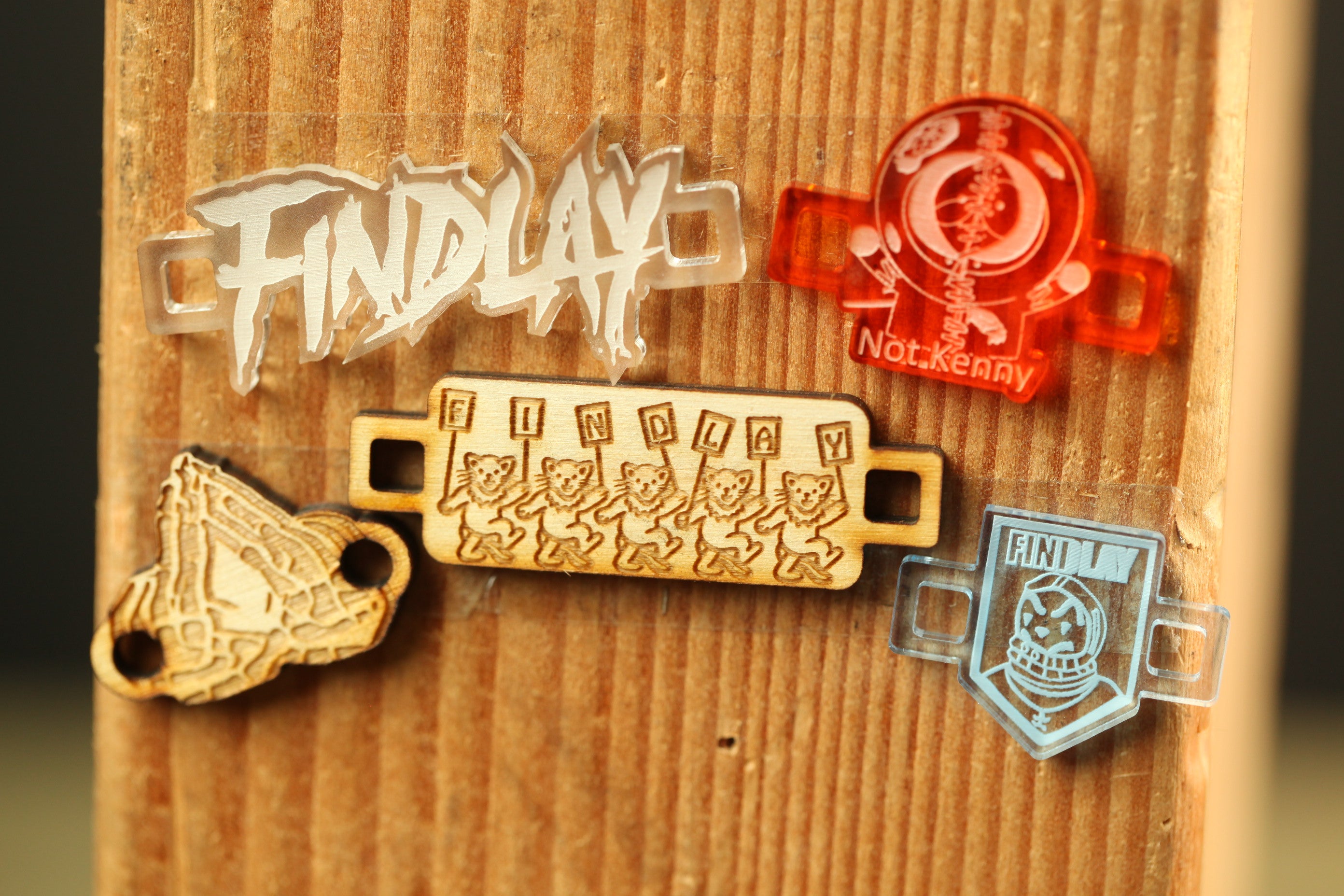 a wooden surface with various printed and engraved items attached to it, including a clear plastic object with the word "FINDlay" and a red object with the text "Not.Kenny".