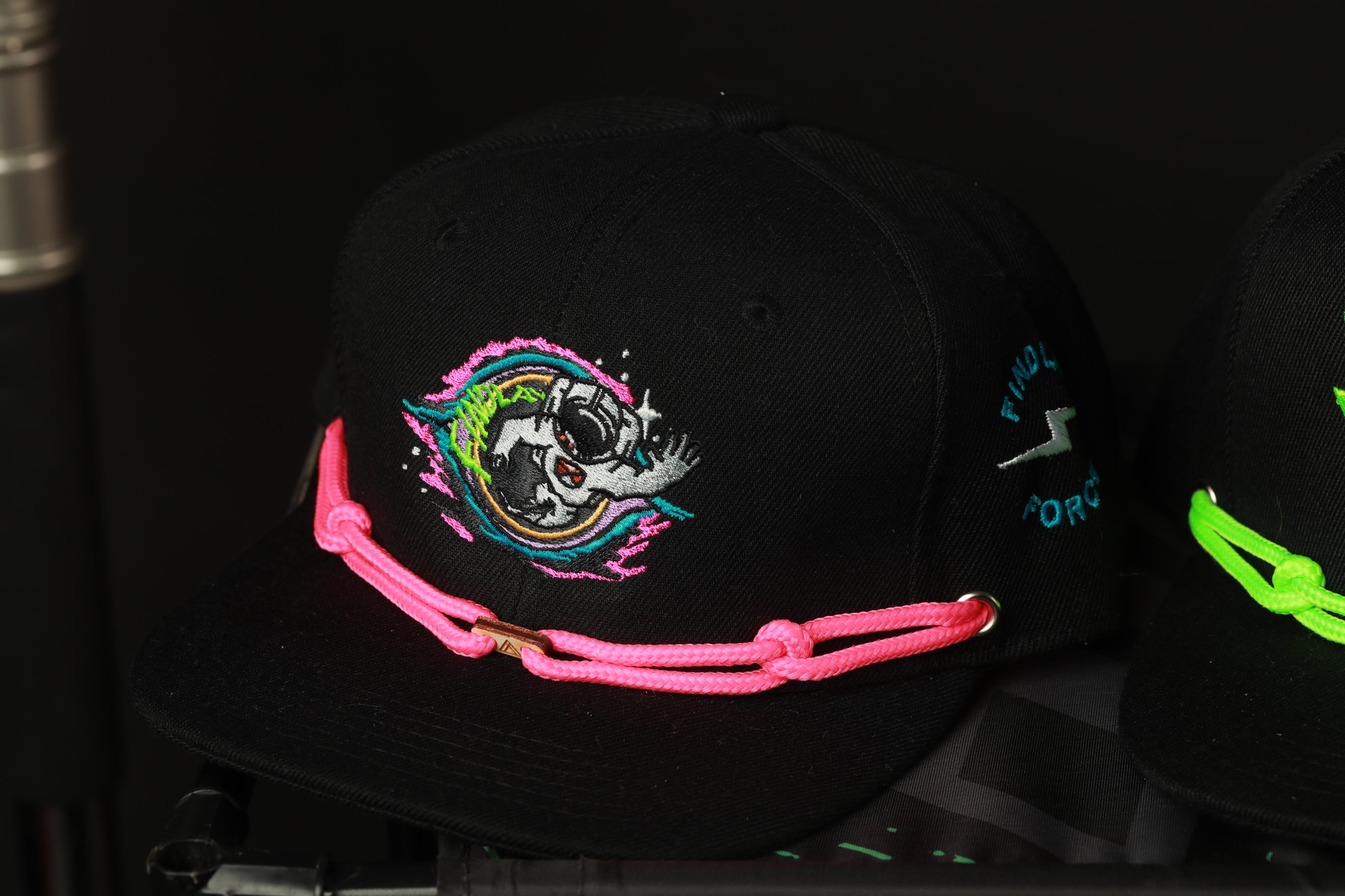 A black baseball cap with a colorful, embroidered design on the front, featuring a skull and various other elements. The cap is adorned with a pink string or chain, adding a pop of color to the overall design.