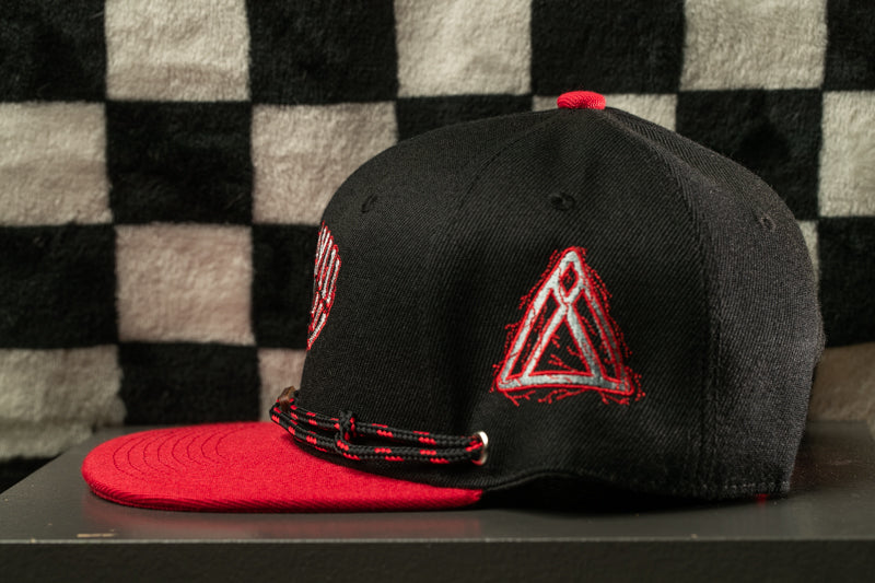 A black baseball cap with a red brim and a red triangle logo on the front, placed on a black surface against a black and white checkered background.