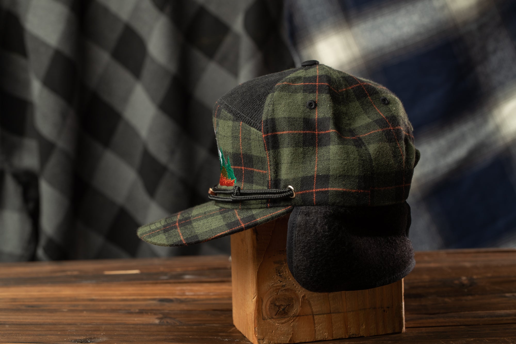 A plaid baseball cap with a black band and a small green patch on the side, displayed on a wooden stand against a blurred background.