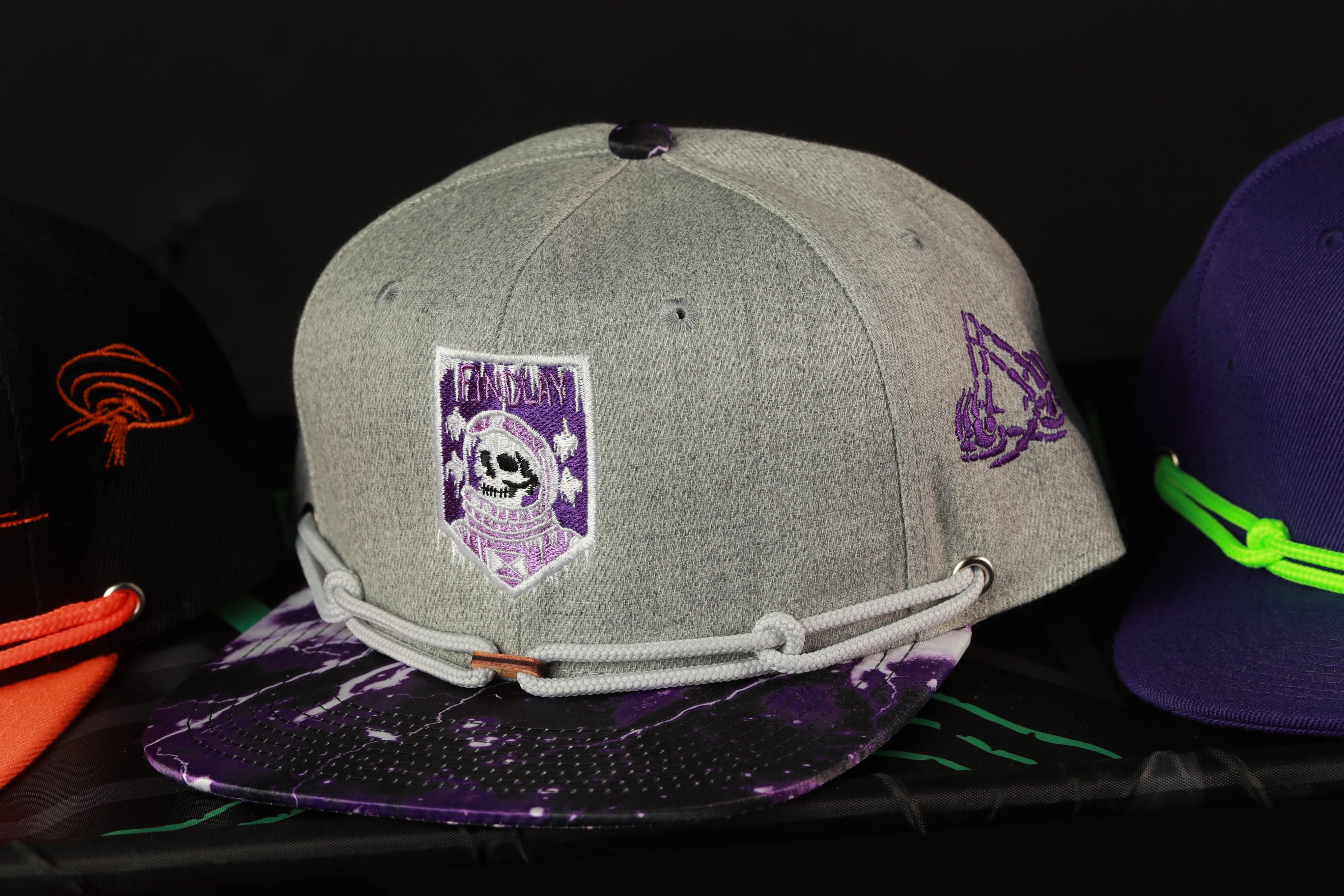 A gray baseball cap with a purple and white design, featuring a skull and crossbones motif, is displayed among other hats on a shelf.