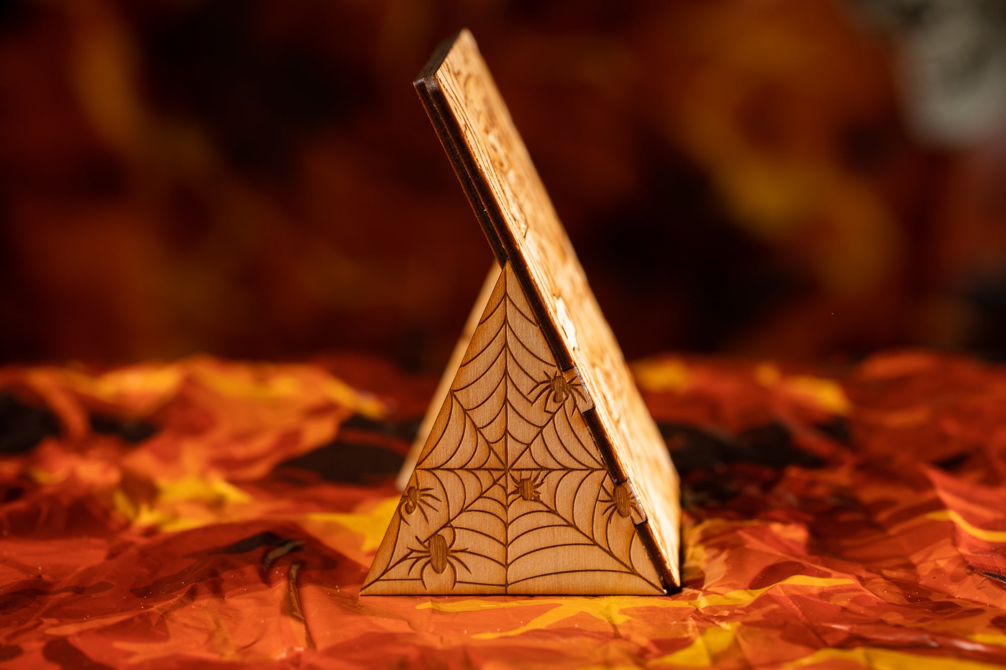 A wooden triangular object with a spider web pattern on it, placed on a surface with orange and yellow autumn leaves.