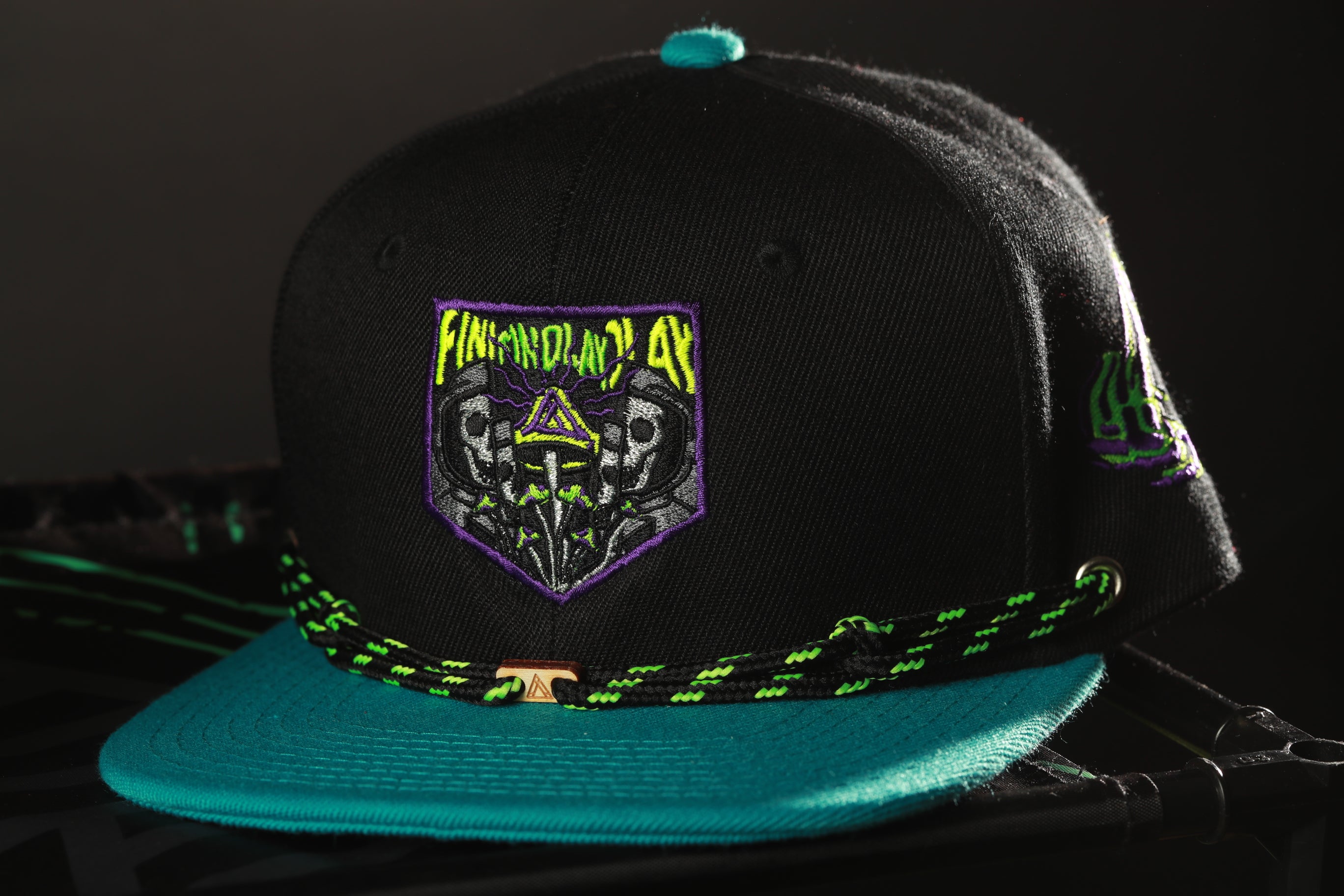 A black baseball cap with a teal brim and a purple and green graphic on the front, featuring a skull and crossbones design.