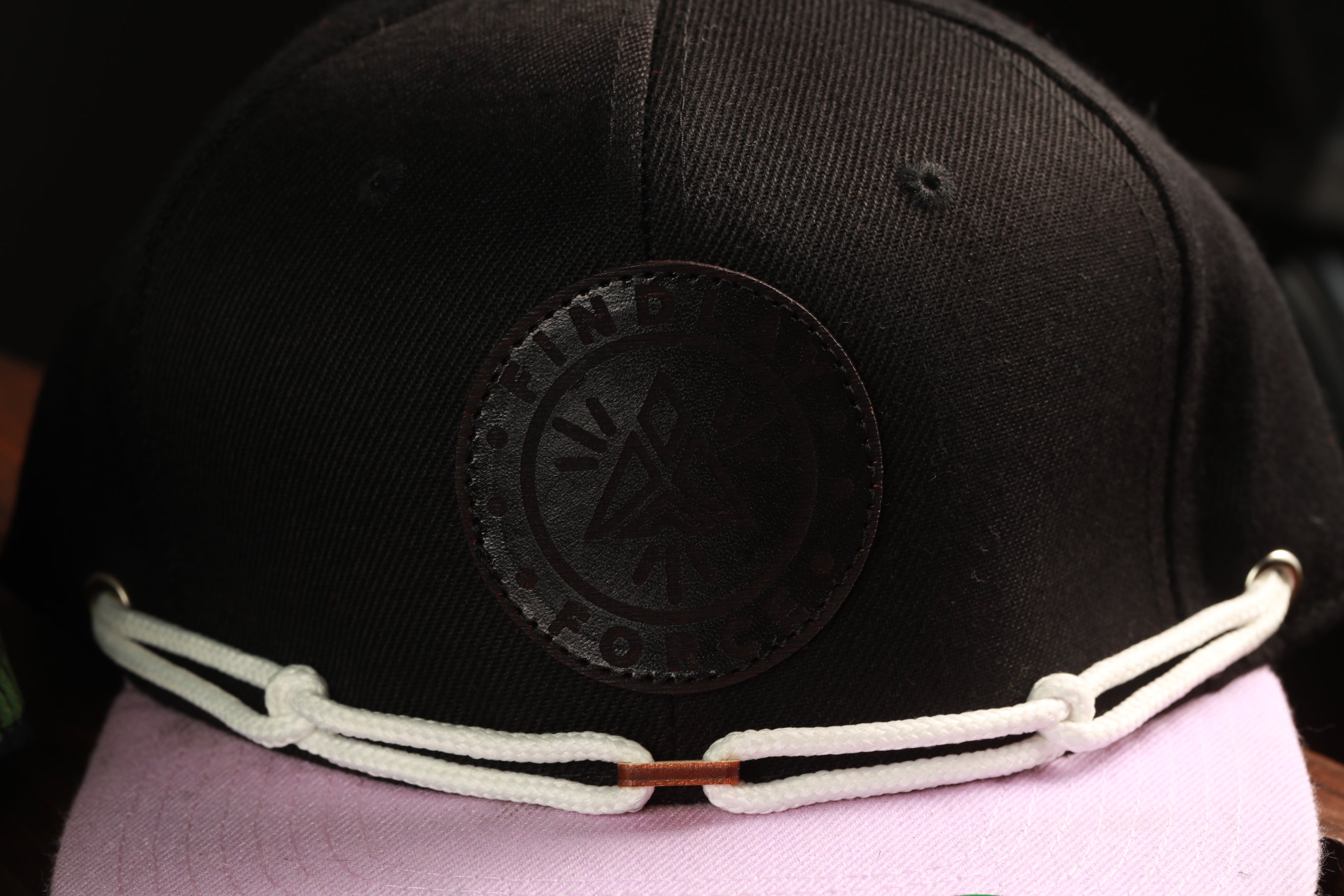 A close-up view of a black baseball cap with a white rope strap and a circular logo on the front.