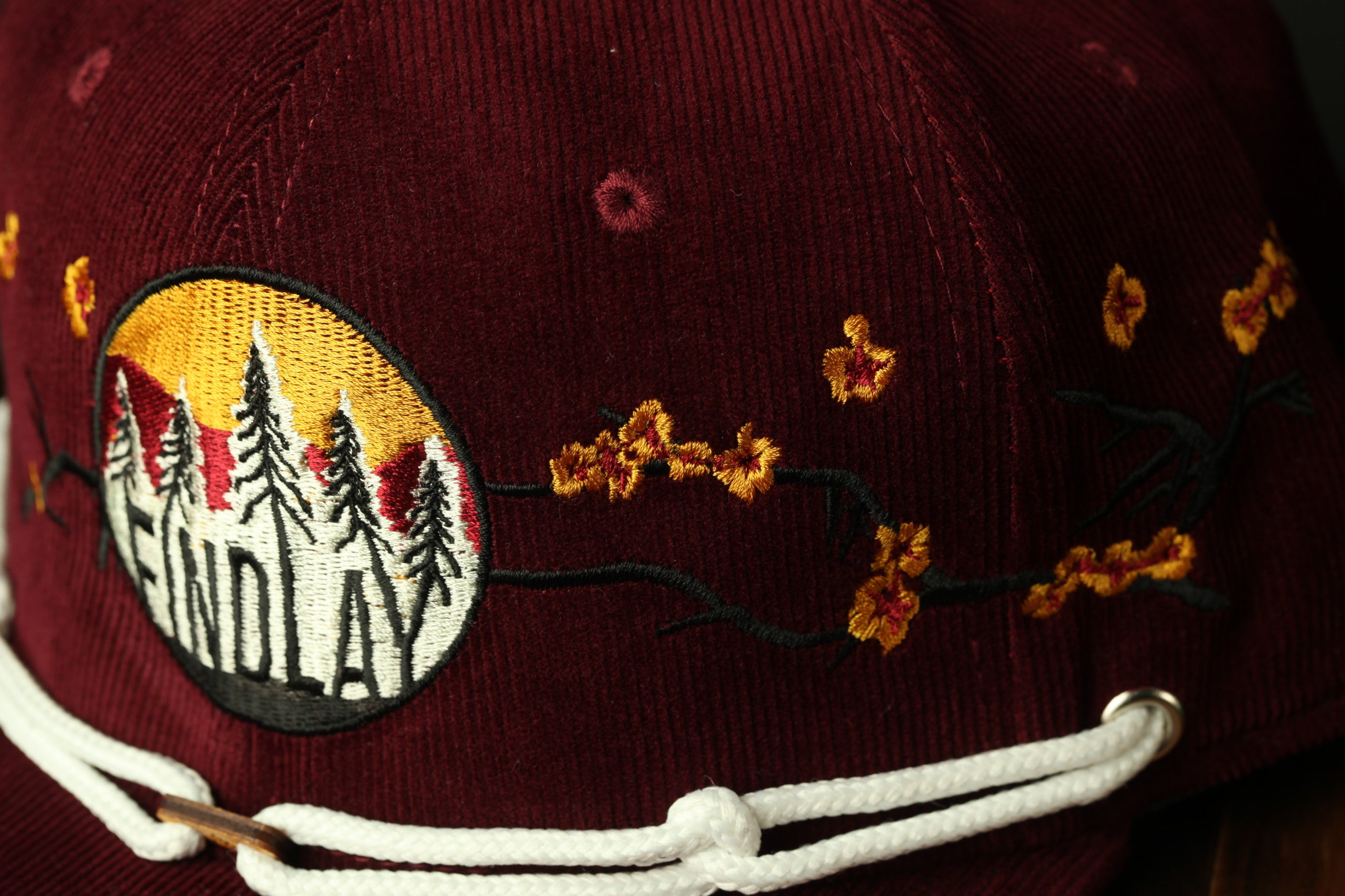 A close-up view of a burgundy baseball cap with a circular embroidered patch featuring a design of trees and flowers.