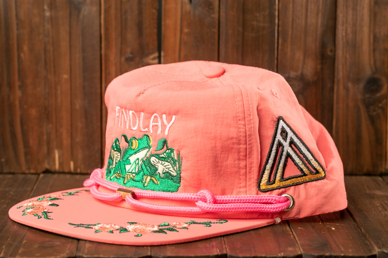 A pink baseball cap with a floral design and the word "FINDLAY" printed on it, sitting on a wooden surface.
