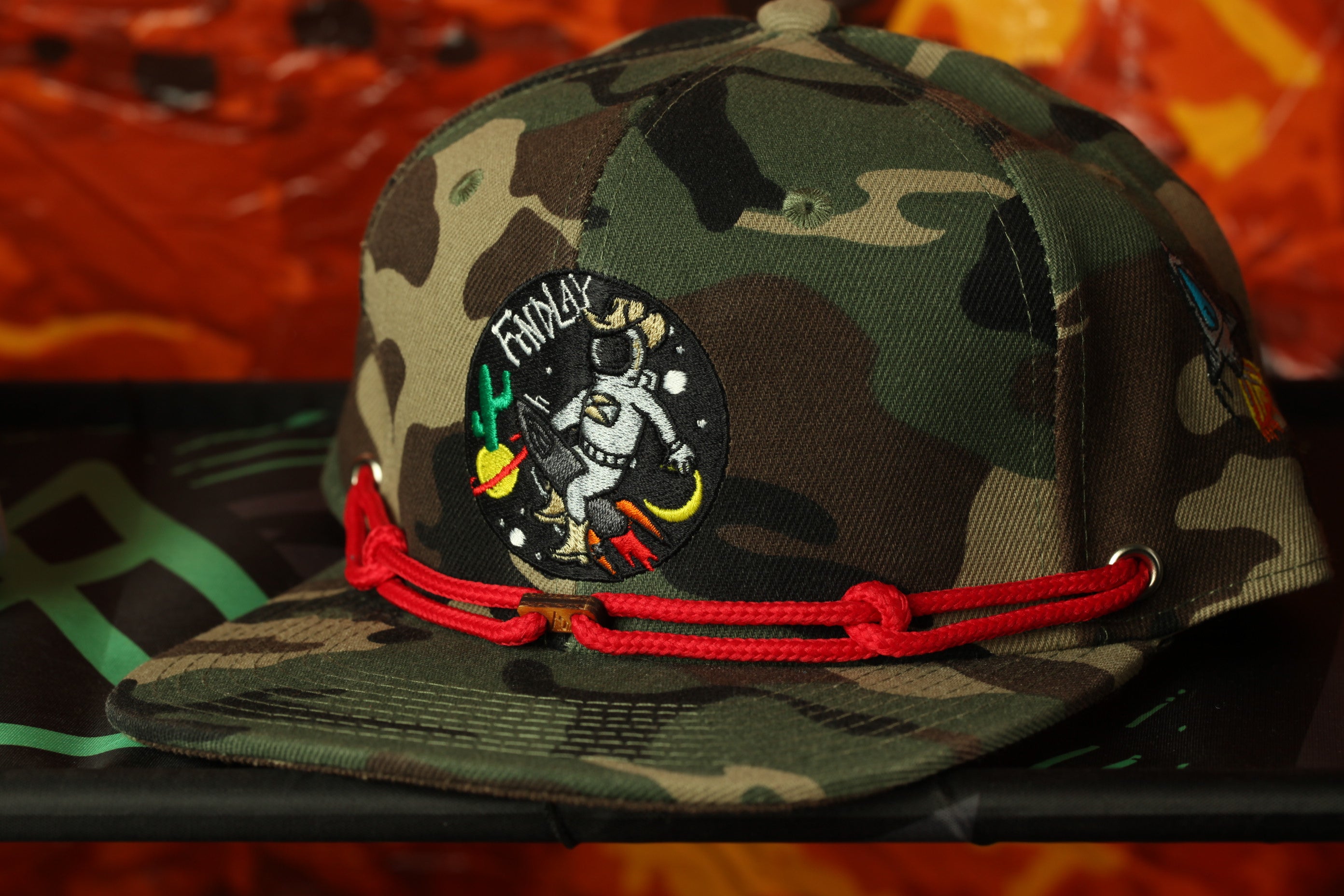 A camouflage baseball cap with a red string and a circular patch featuring a cartoon character is displayed against a red and orange background.