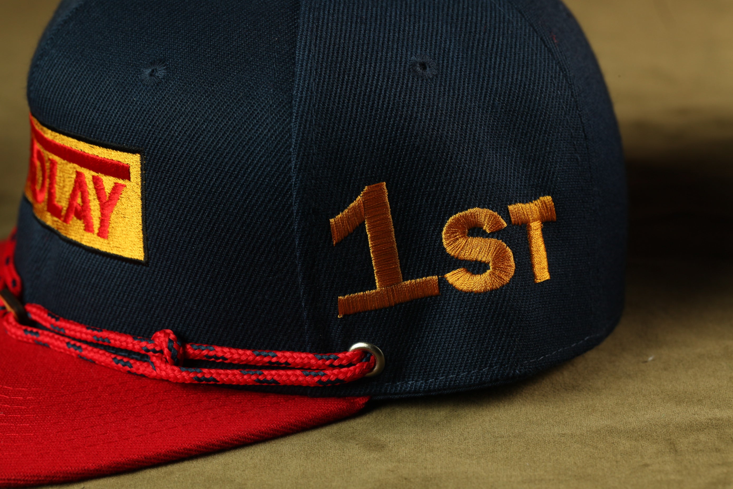 A close-up view of a black baseball cap with red and gold embroidered details, including the text "1ST" on the side.