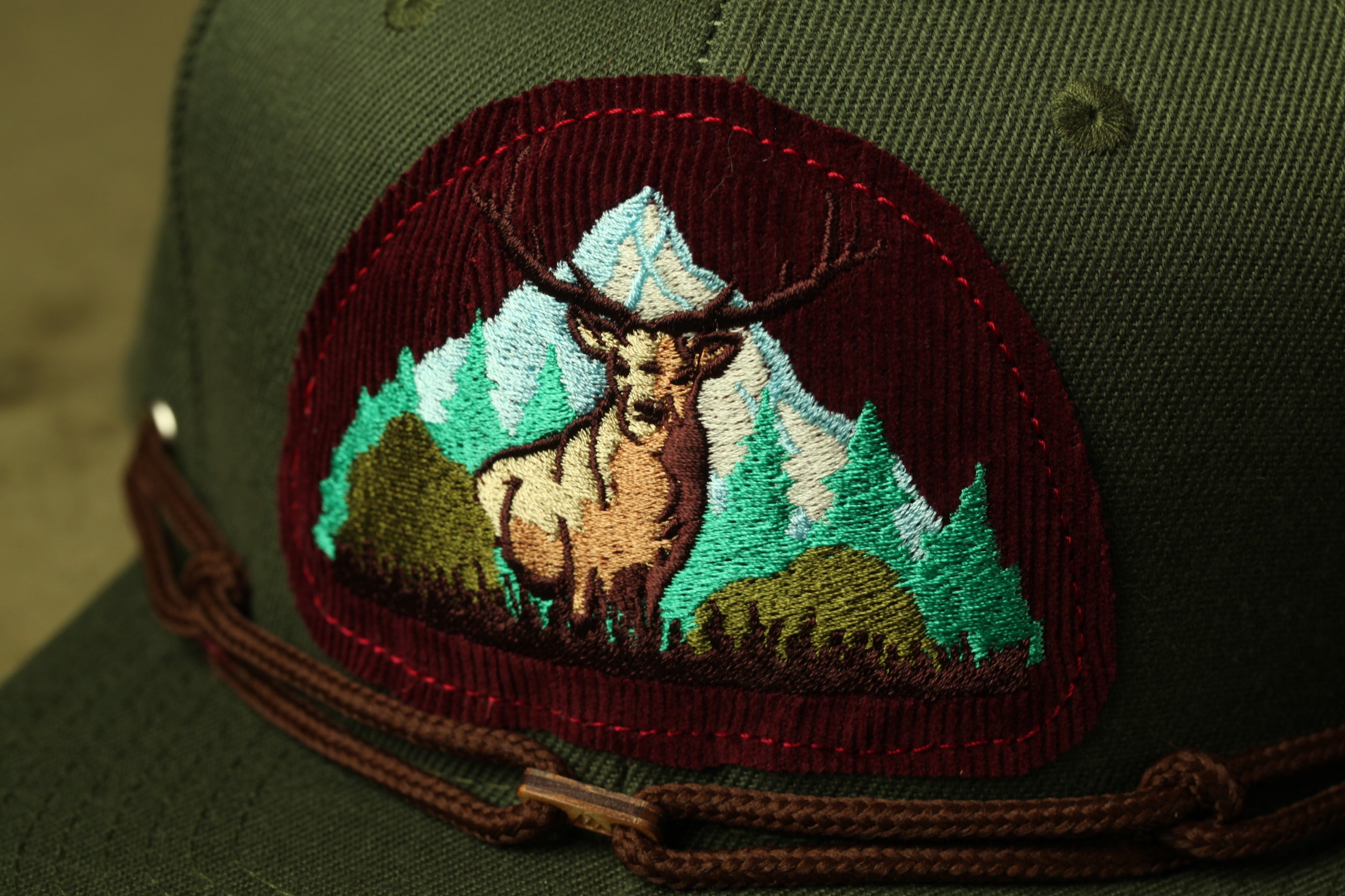 A close-up of a green baseball cap with a patch featuring a deer standing in front of a mountain landscape.