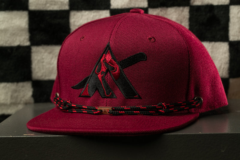 A maroon baseball cap with a black logo and a black and red braided strap is displayed against a black and white checkered background.