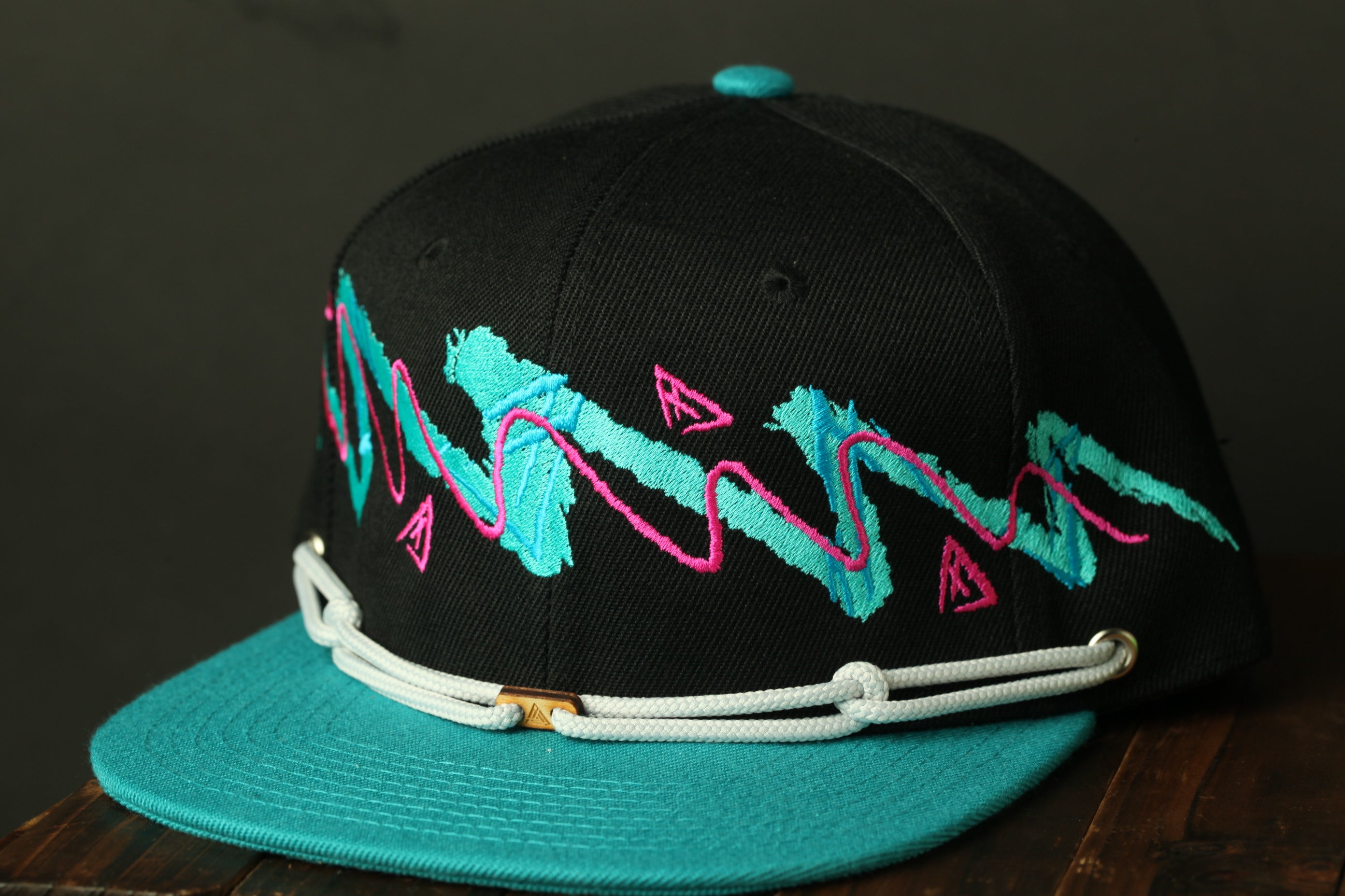 A black baseball cap with a teal brim and colorful embroidered designs on the front.